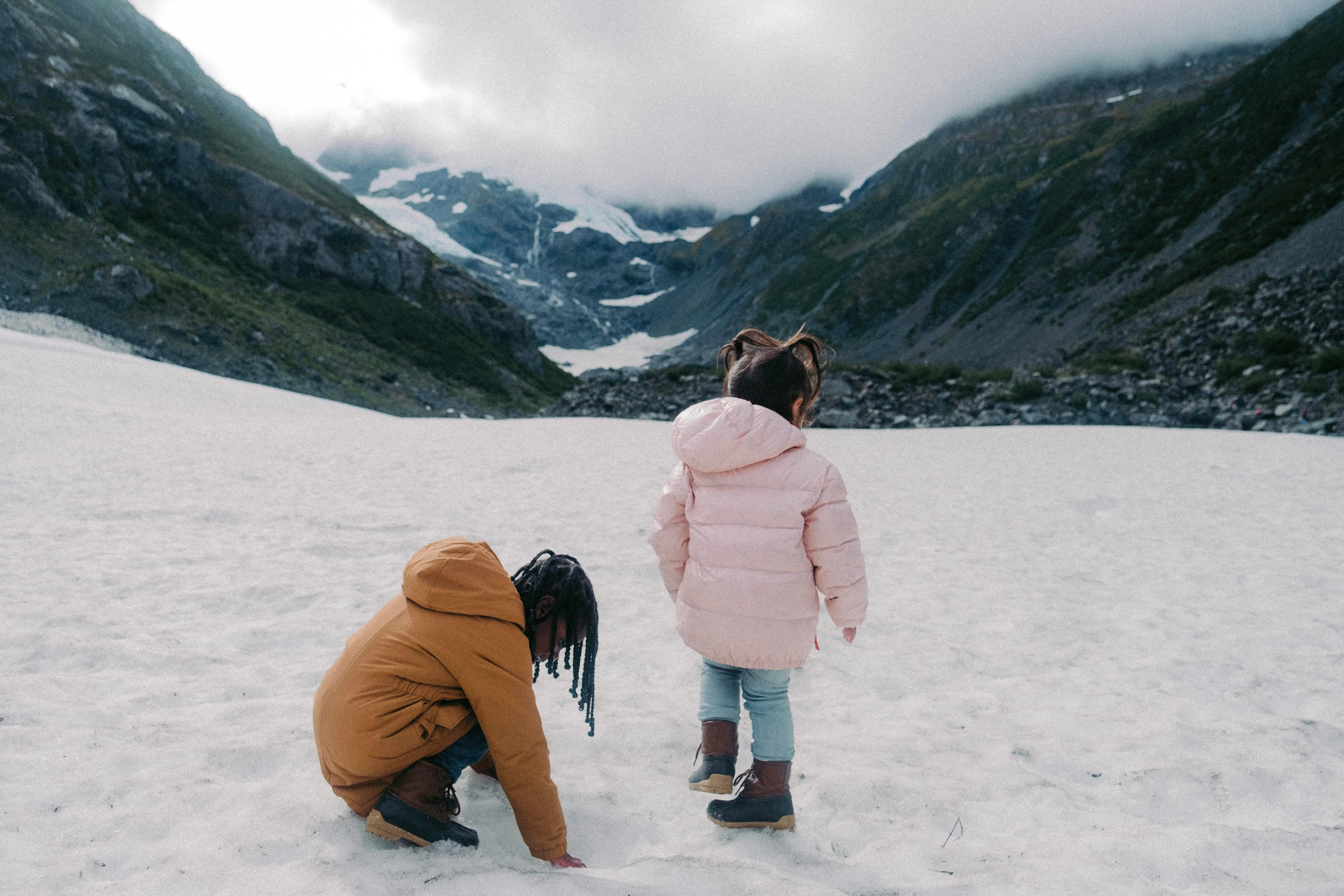 Zwei Kinder in Winterkleidung stehen auf einer Schneefläche vor bewölkten Bergen.