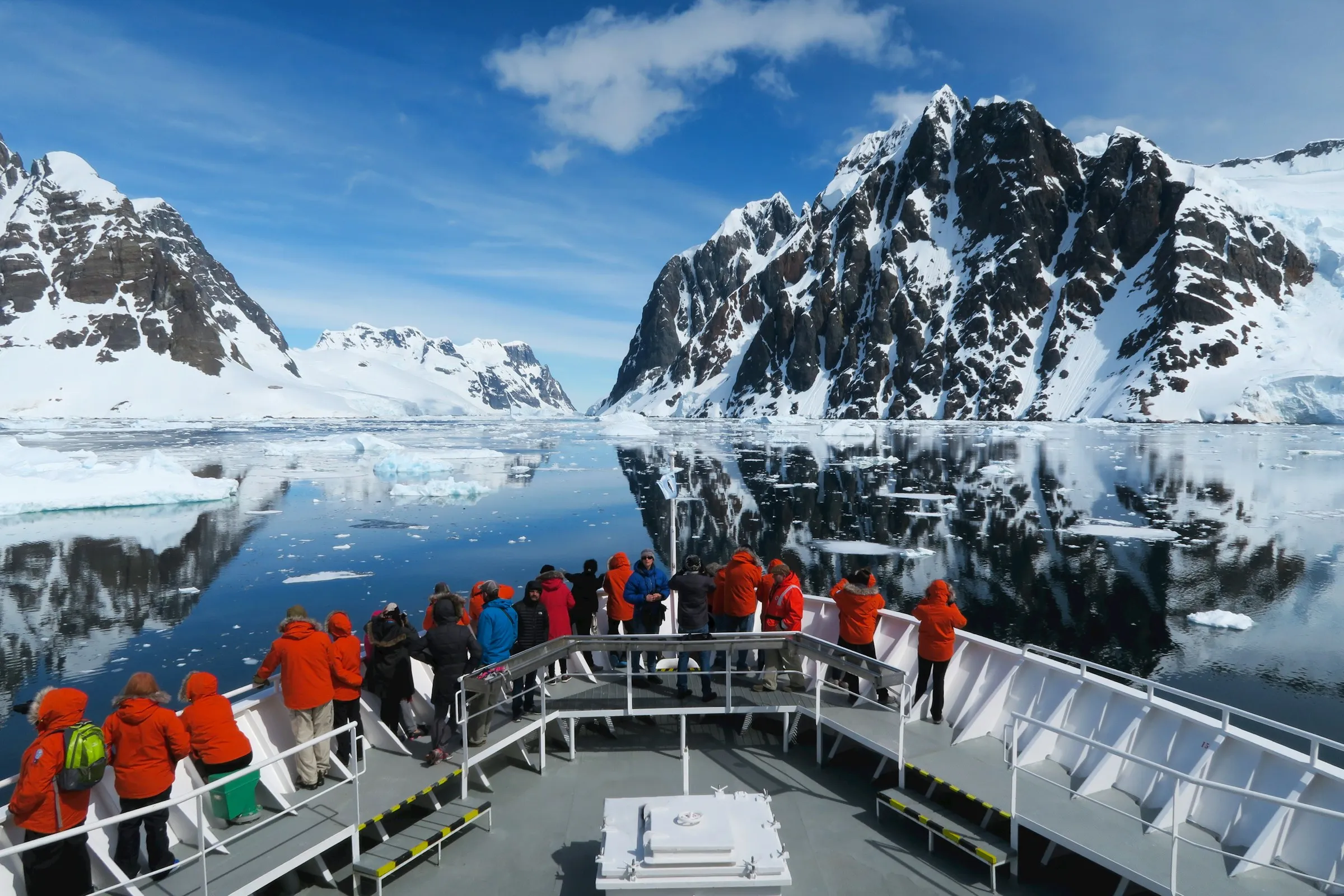 Gruppe von Menschen in orangen Jacken auf dem Deck eines Schiffes vor schneebedeckten Bergen und spiegelglattem Wasser mit treibenden Eisschollen.