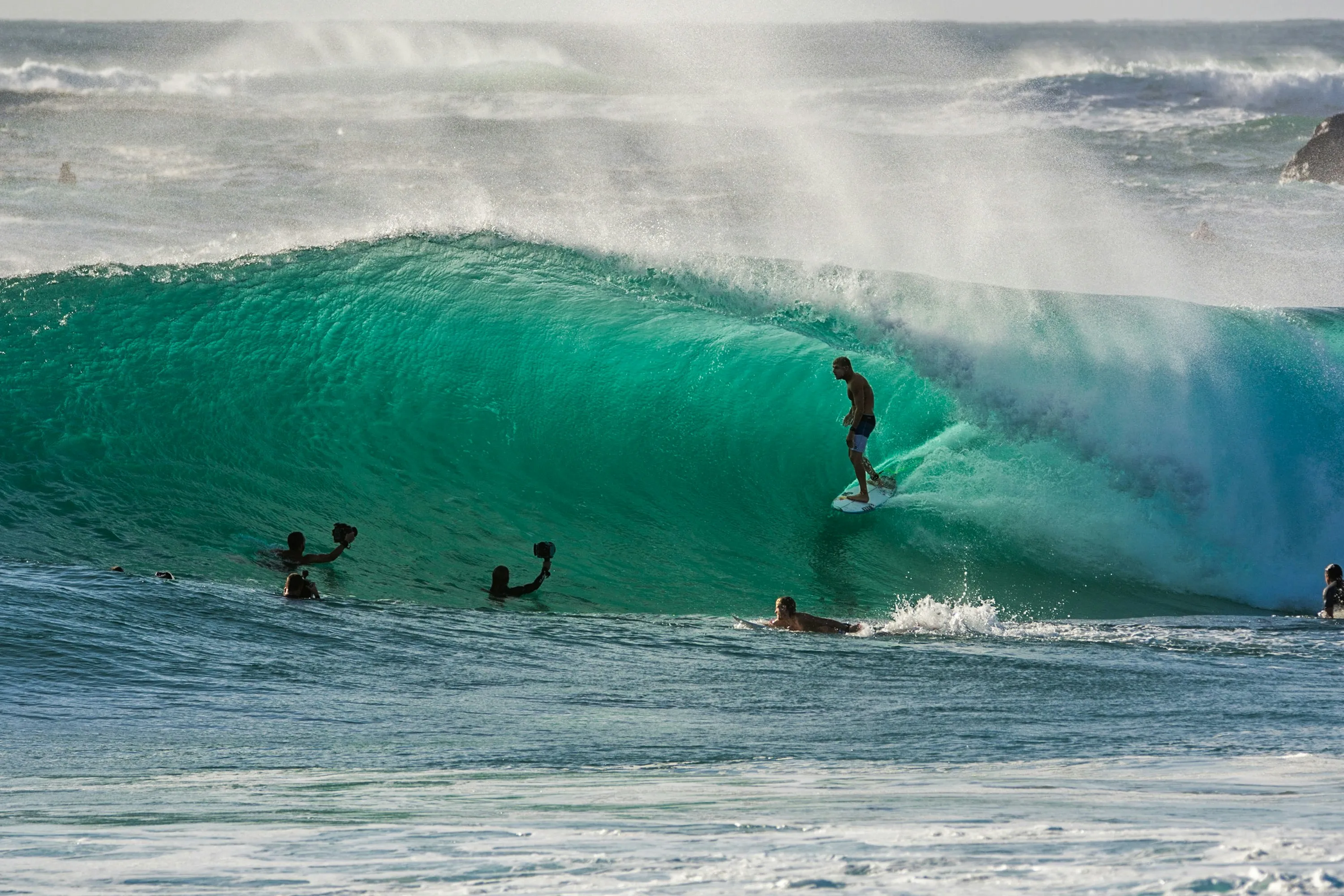 Surfer steht auf einem Surfbrett in einer großen Welle, mehrere weitere Personen sind im Wasser in der Nähe.