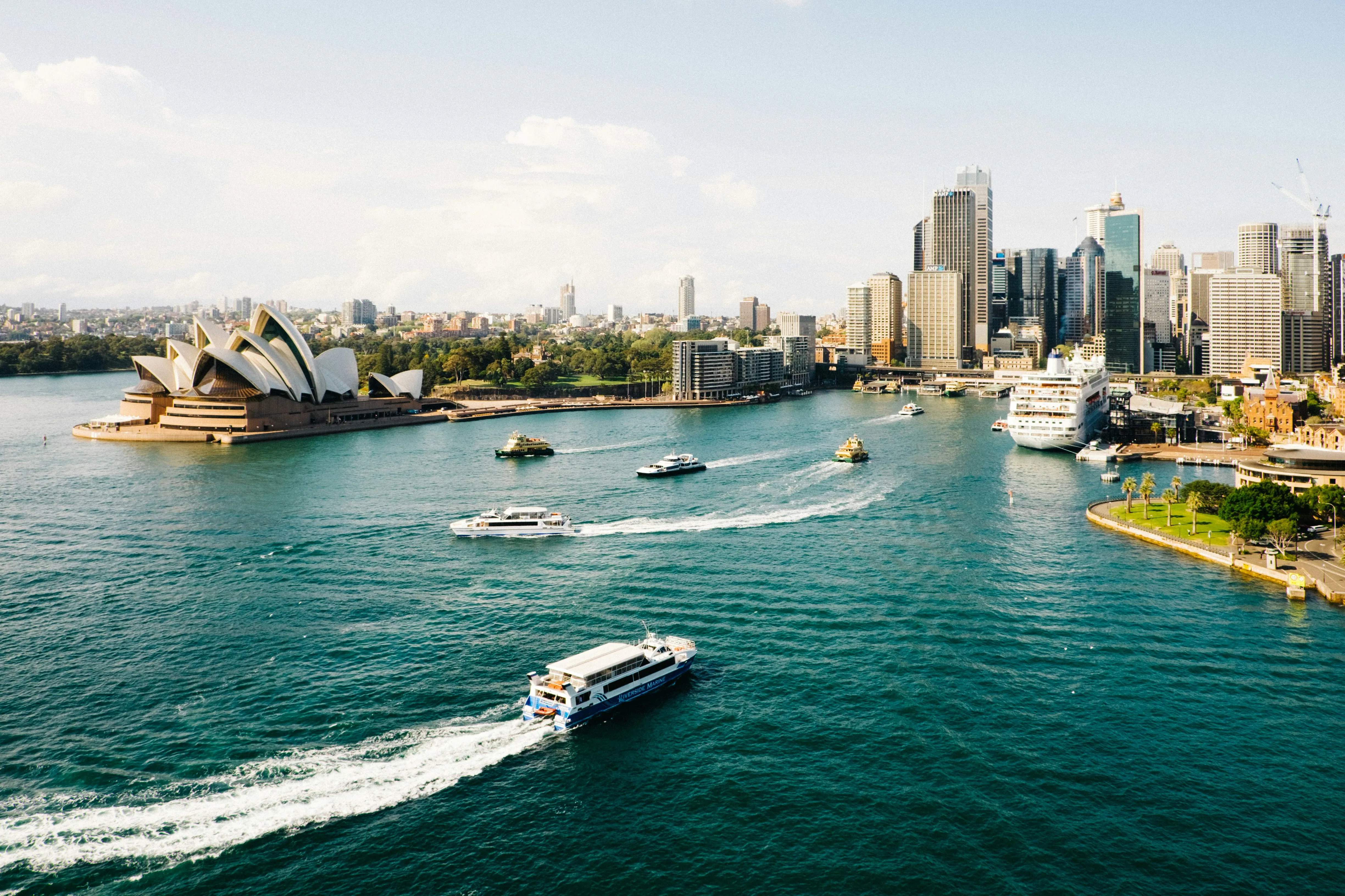 Blick auf die Sydneyer Hafenbucht mit dem Opernhaus links, mehreren Fähren auf dem Wasser und einer Skyline aus Hochhäusern im Hintergrund.