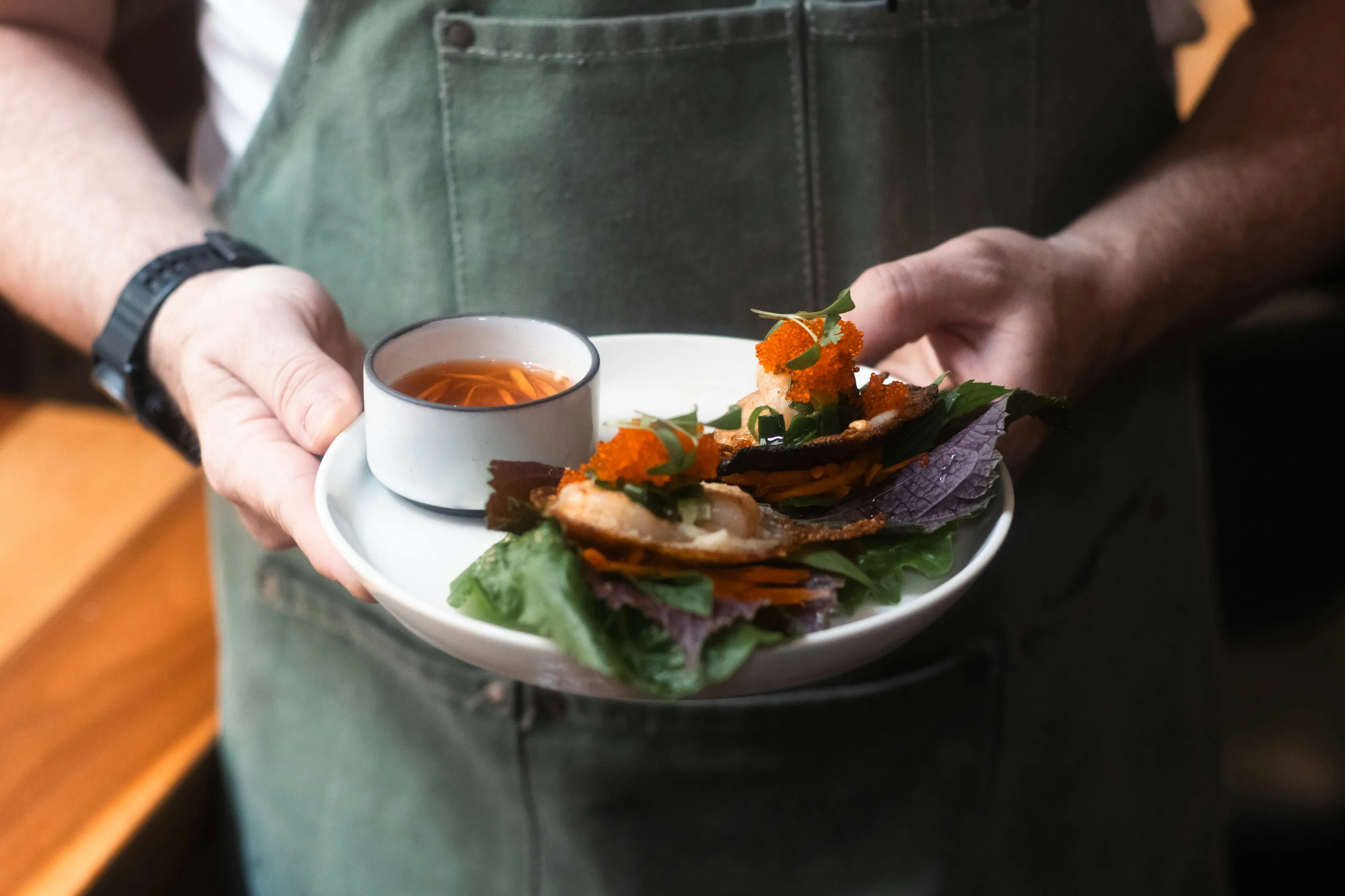 Person hält Teller mit zwei kleinen Tacos auf Salatblättern und einer kleinen Schale mit roter Sauce.