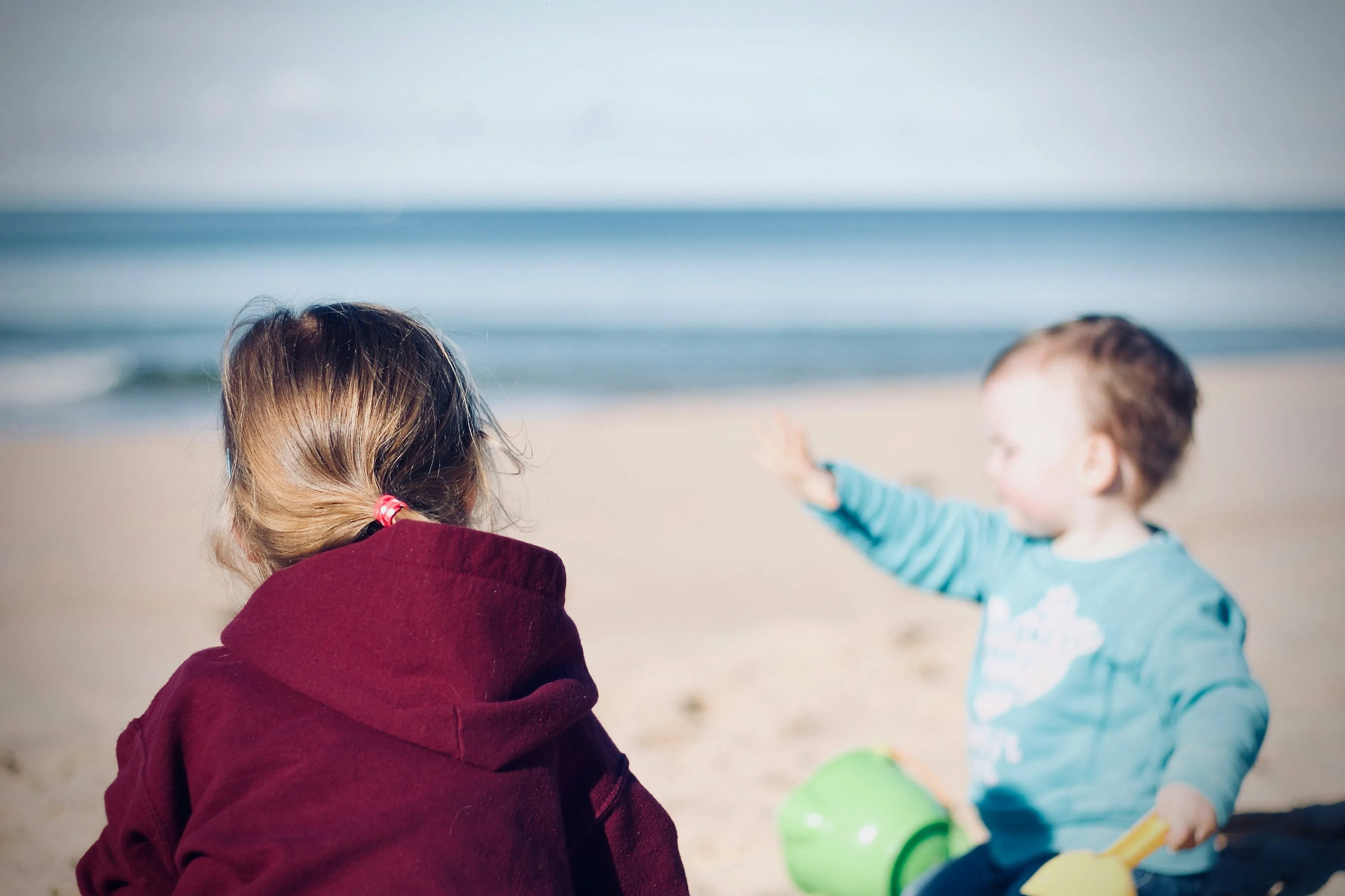 Zwei Kinder sitzen am Strand, eines trägt einen dunkelroten Kapuzenpullover, das andere ein hellblaues Oberteil und hält einen grünen Eimer.