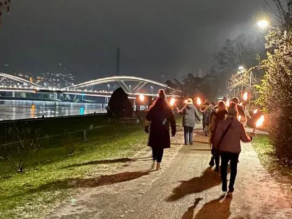 Gruppe von vier Personen sitzt auf einer Wiese mit Blick auf einen See und bewaldete Hügel unter blauem Himmel.
