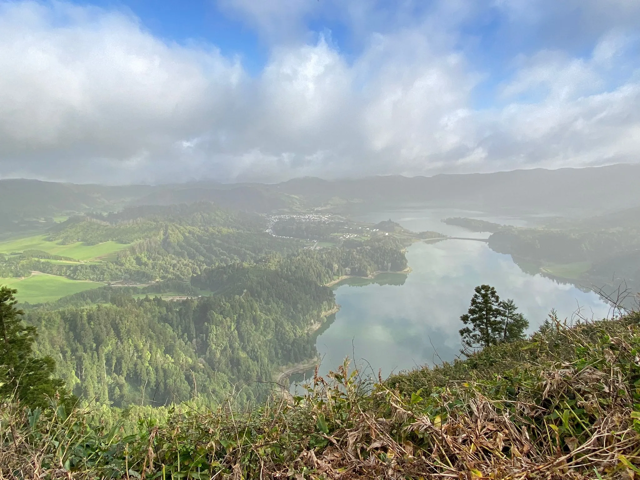 Blick von einem Hügel auf einen See, umgeben von bewaldeten Hügeln und grünen Wiesen unter bewölktem Himmel.