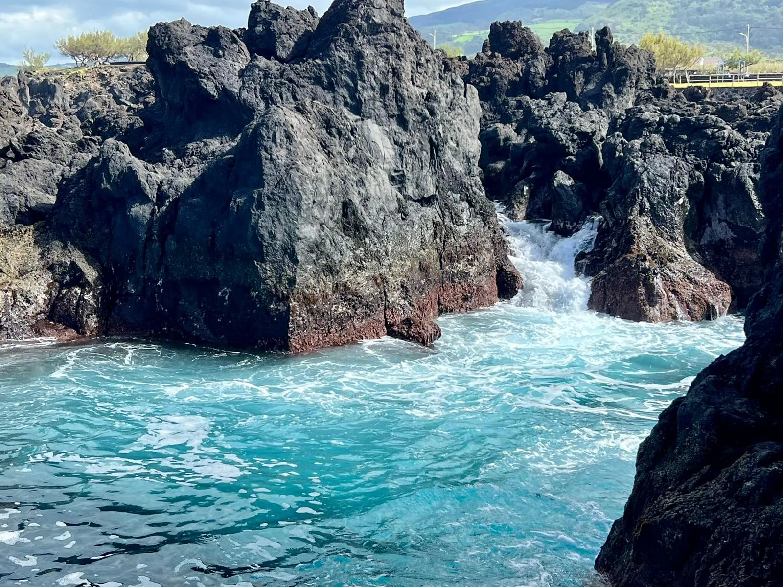 Felsige Küste mit schwarzem Vulkangestein und türkisfarbenem Wasser, das gegen die Felsen schlägt.
