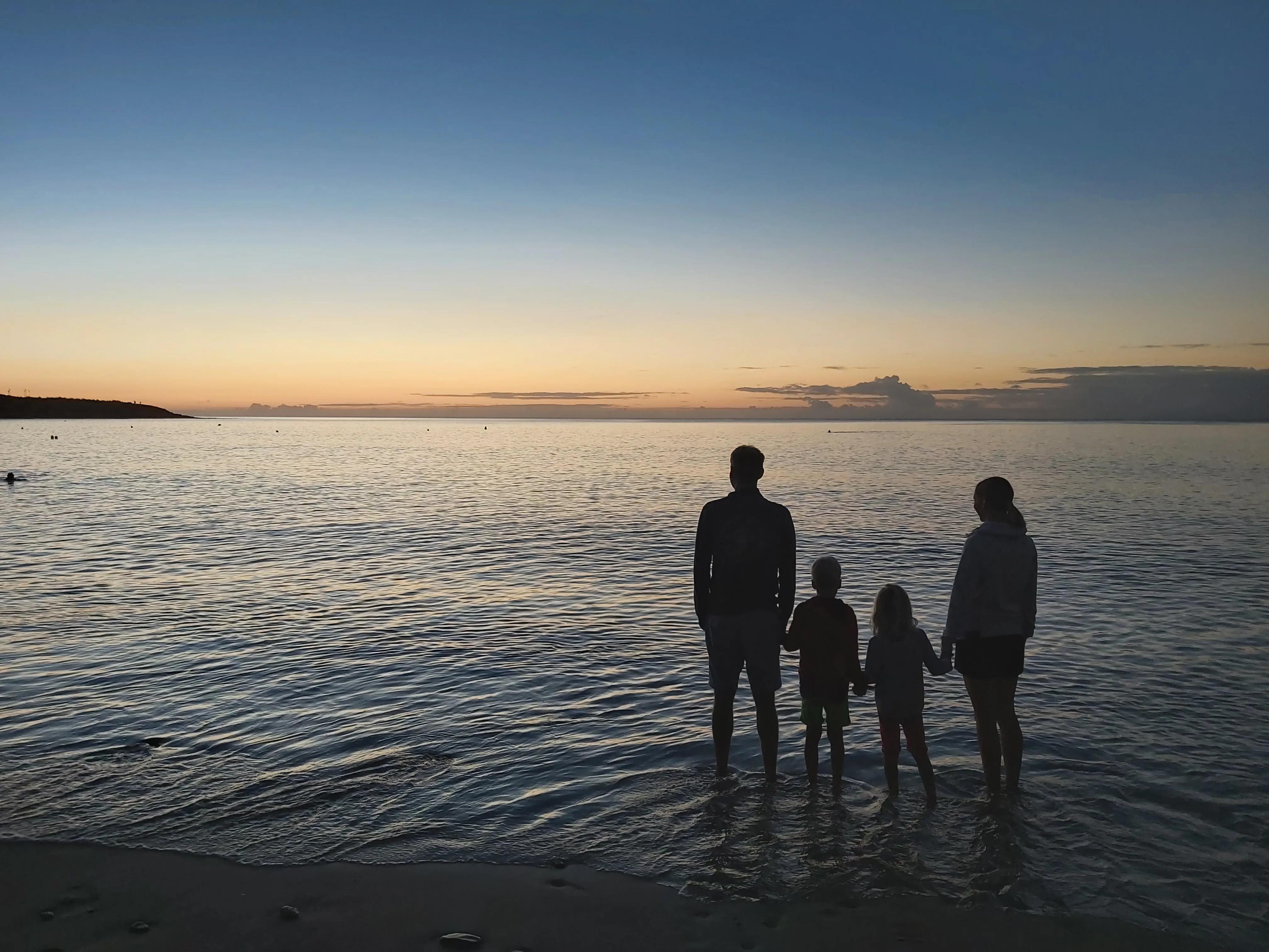 Silhouetten von zwei Erwachsenen und zwei Kindern stehen im flachen Wasser am Strand bei Sonnenuntergang.