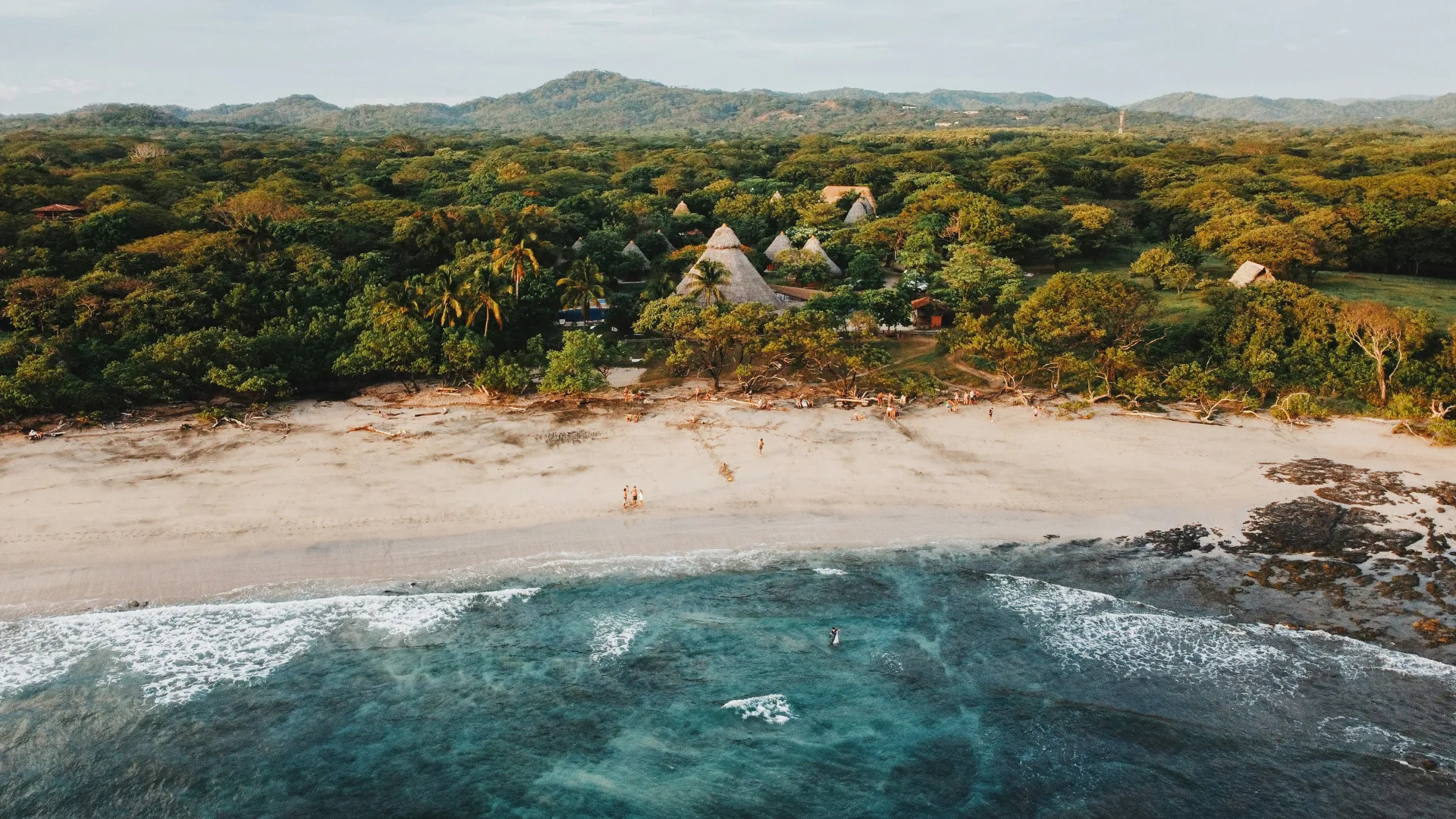 Luftaufnahme eines Strandes mit Wellen, dahinter ein bewaldetes Gebiet mit mehreren spitz zulaufenden, strohgedeckten Hütten und Hügeln im Hintergrund.