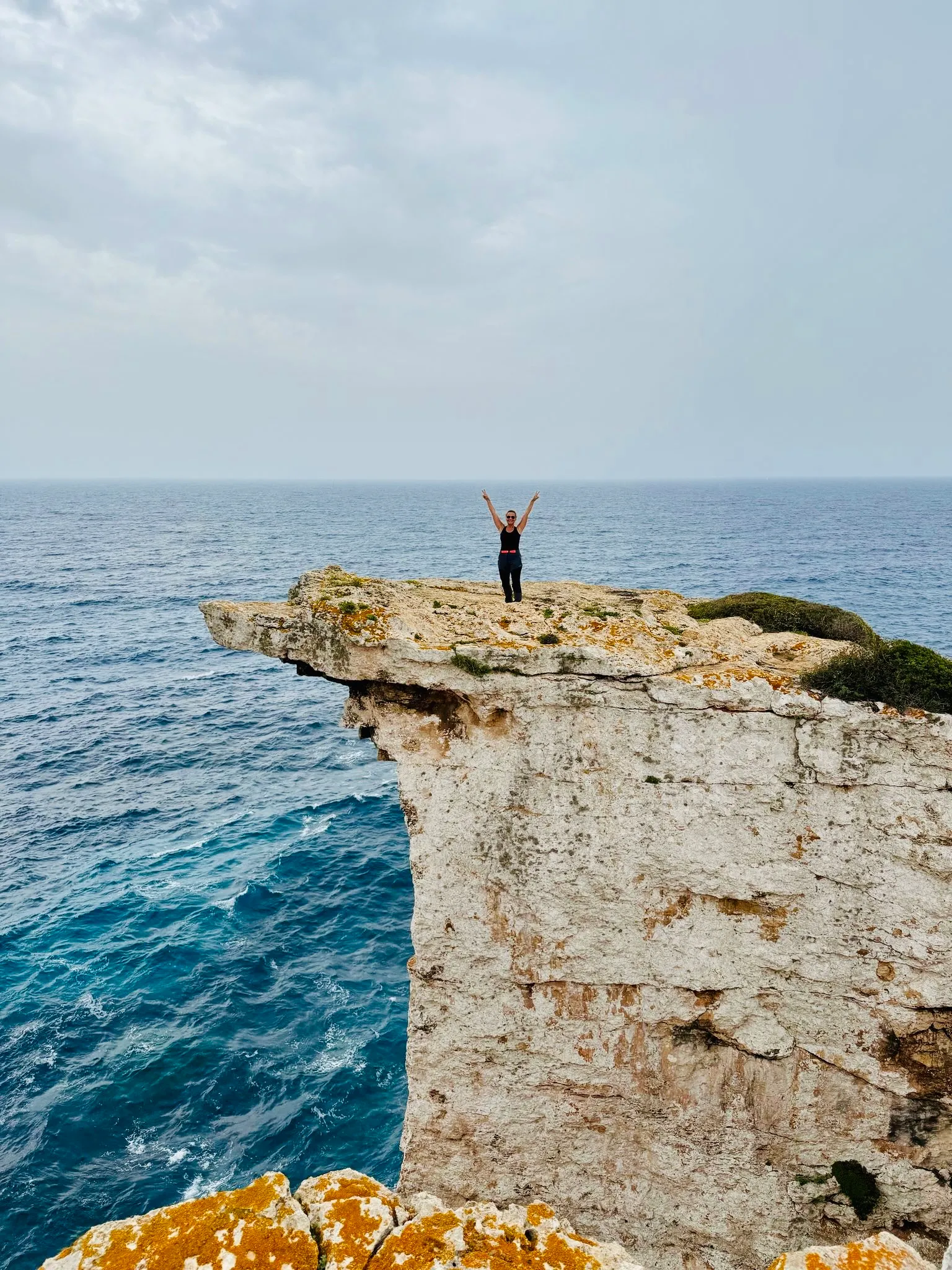 Person steht mit erhobenen Armen auf einer Klippe über dem Meer auf Mallorca, bewölkter Himmel im Hintergrund.