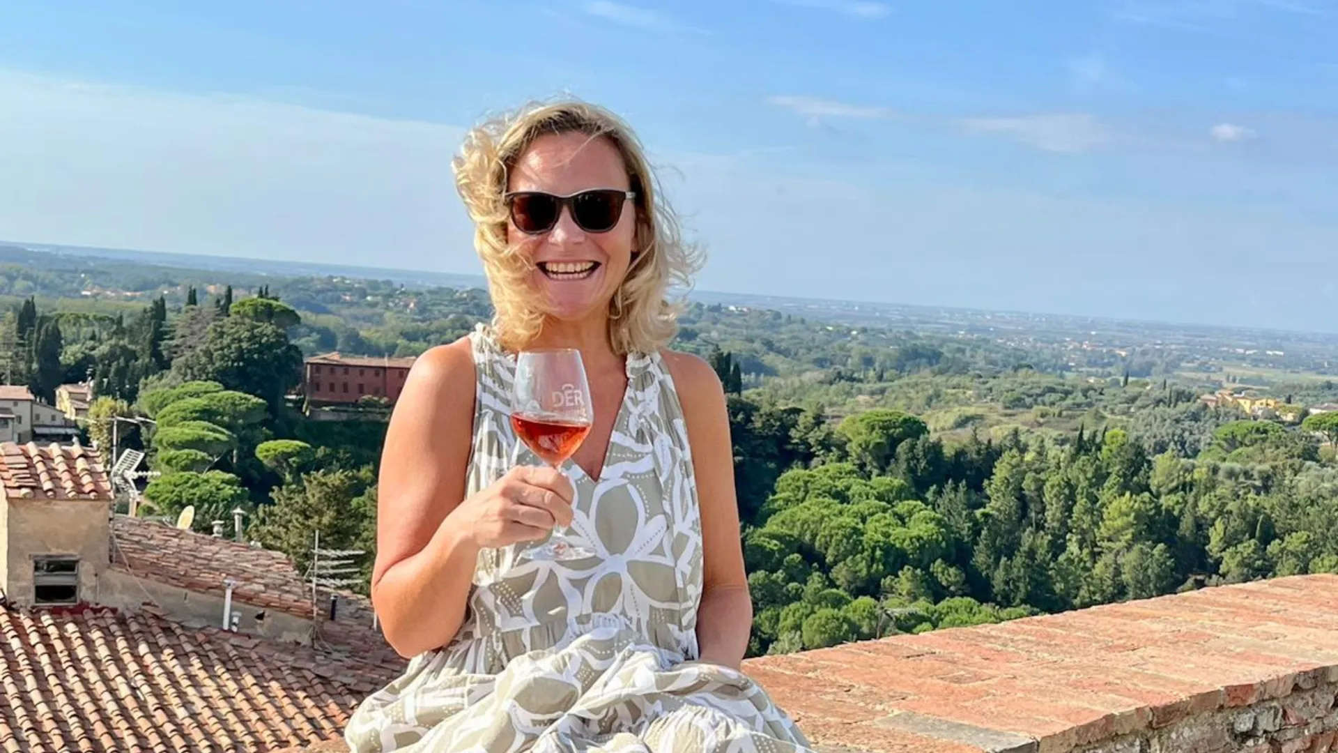 Frau in weiß-grau gemustertem Kleid sitzt auf einer Ziegelmauer und hält ein Glas Roséwein, im Hintergrund Landschaft mit Bäumen und Gebäuden.