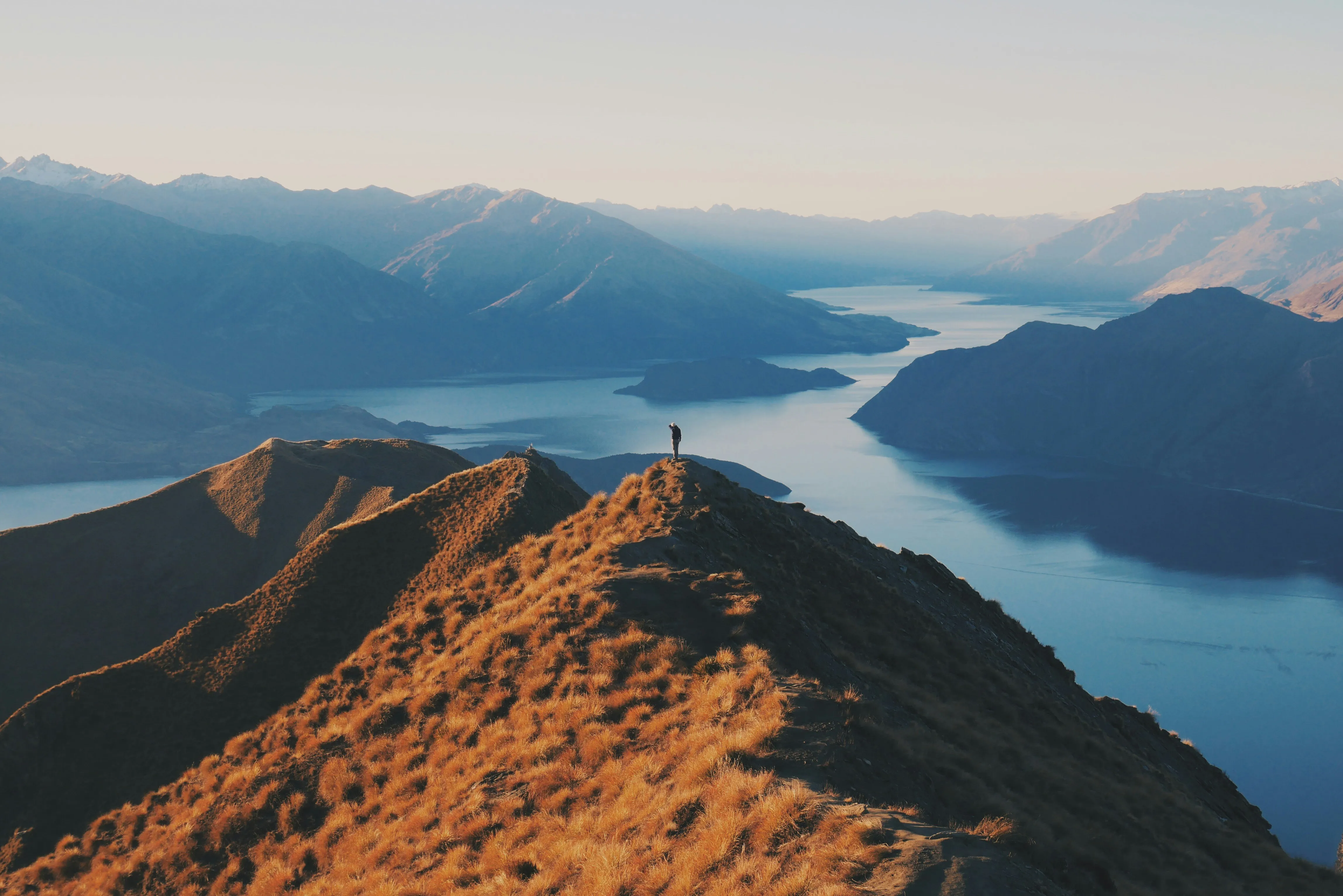Person steht auf einem grasbewachsenen Bergkamm mit Blick auf einen See und dahinter liegende Bergketten.