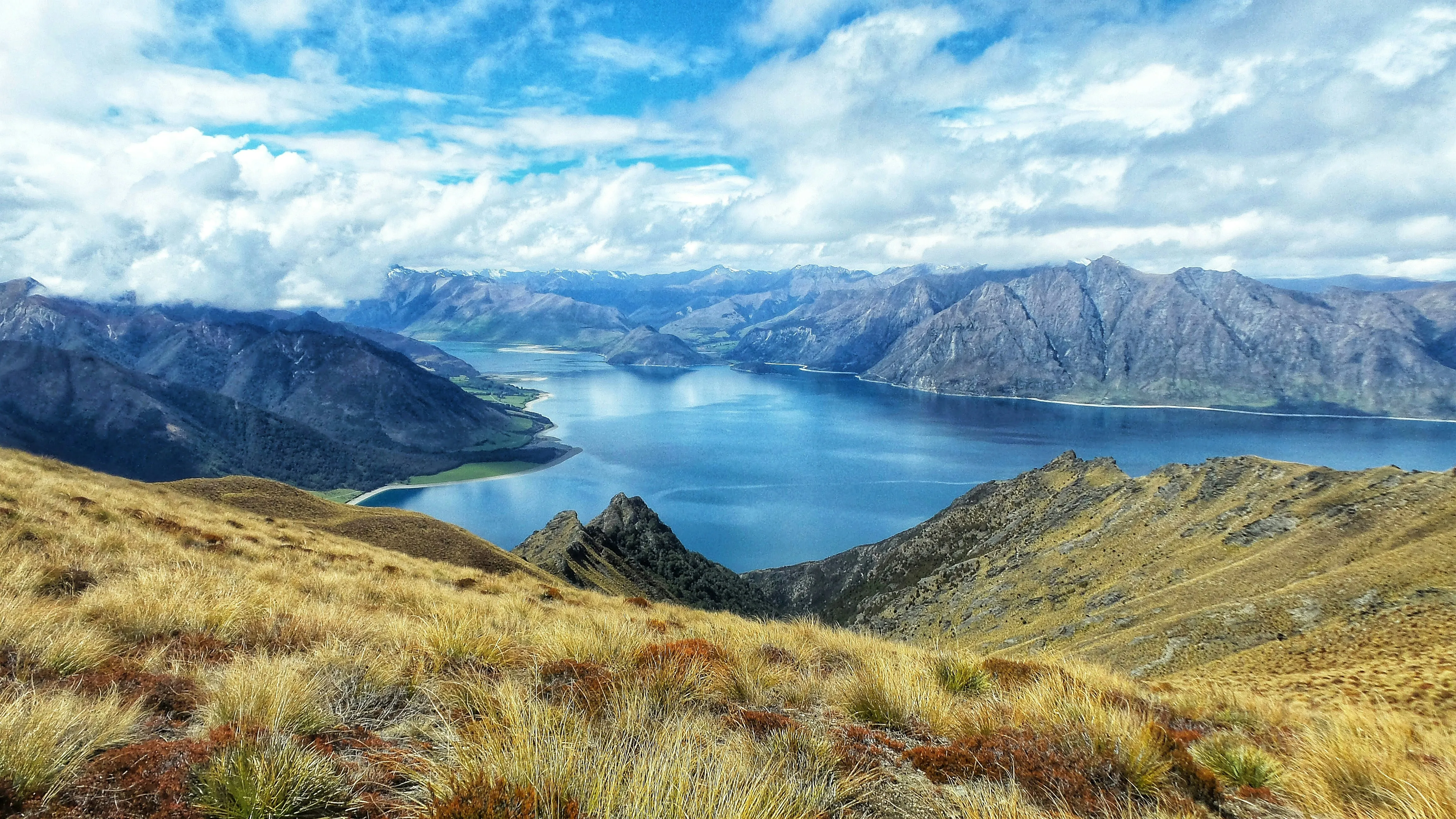 Berglandschaft mit See in der Mitte, umgeben von bewachsenen Hügeln und Bergen unter bewölktem Himmel.
