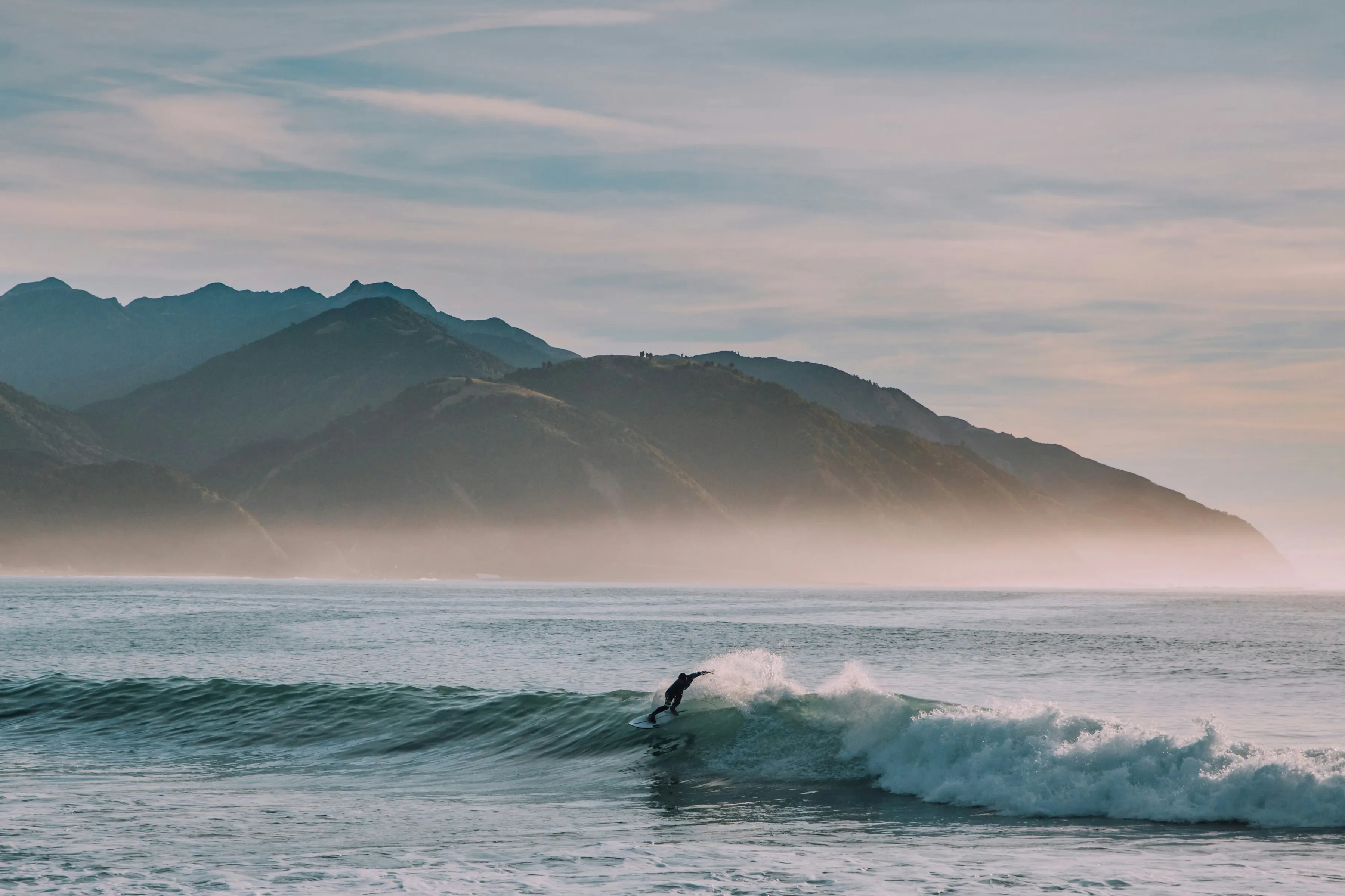 Surfer auf einer Welle vor einer Bergkette im Dunst am Meer.