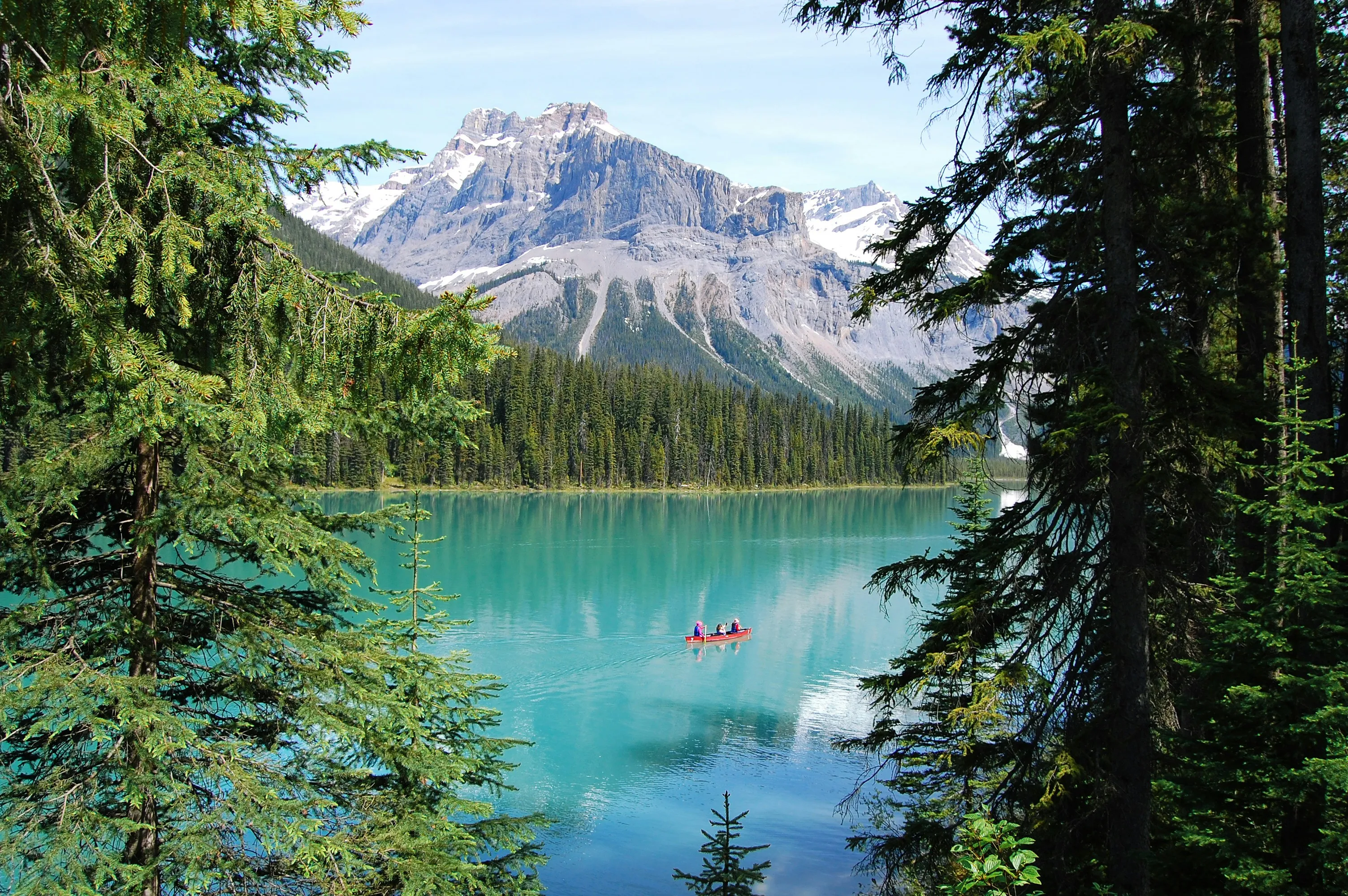 Zwei Personen paddeln in einem Kanu auf einem türkisfarbenen Bergsee, umgeben von Nadelbäumen und Bergen im Hintergrund.