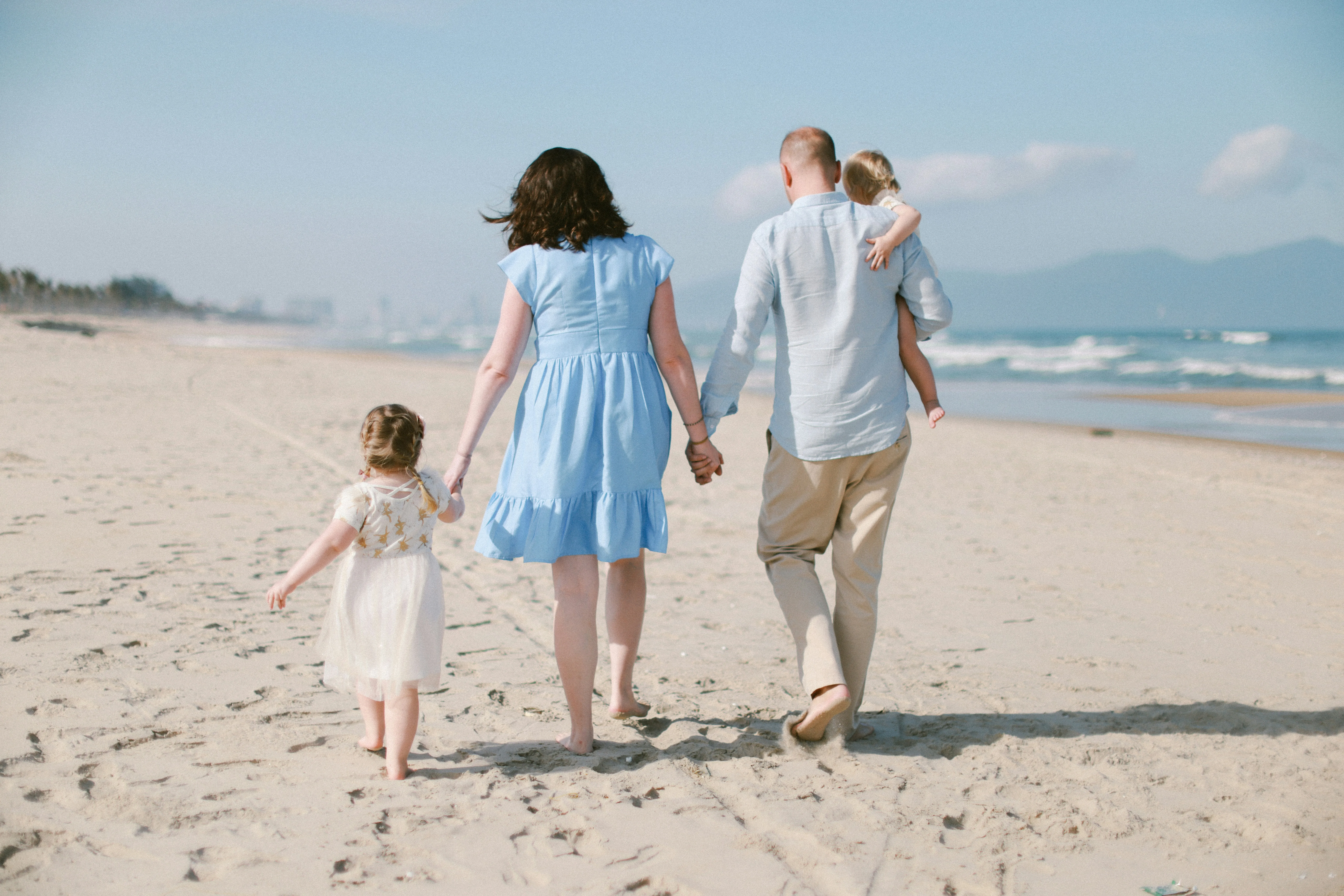Eine Familie mit zwei Kindern geht barfuß am Strand entlang, die Erwachsenen halten jeweils ein Kind an der Hand, der Mann trägt ein Kind auf dem Arm.