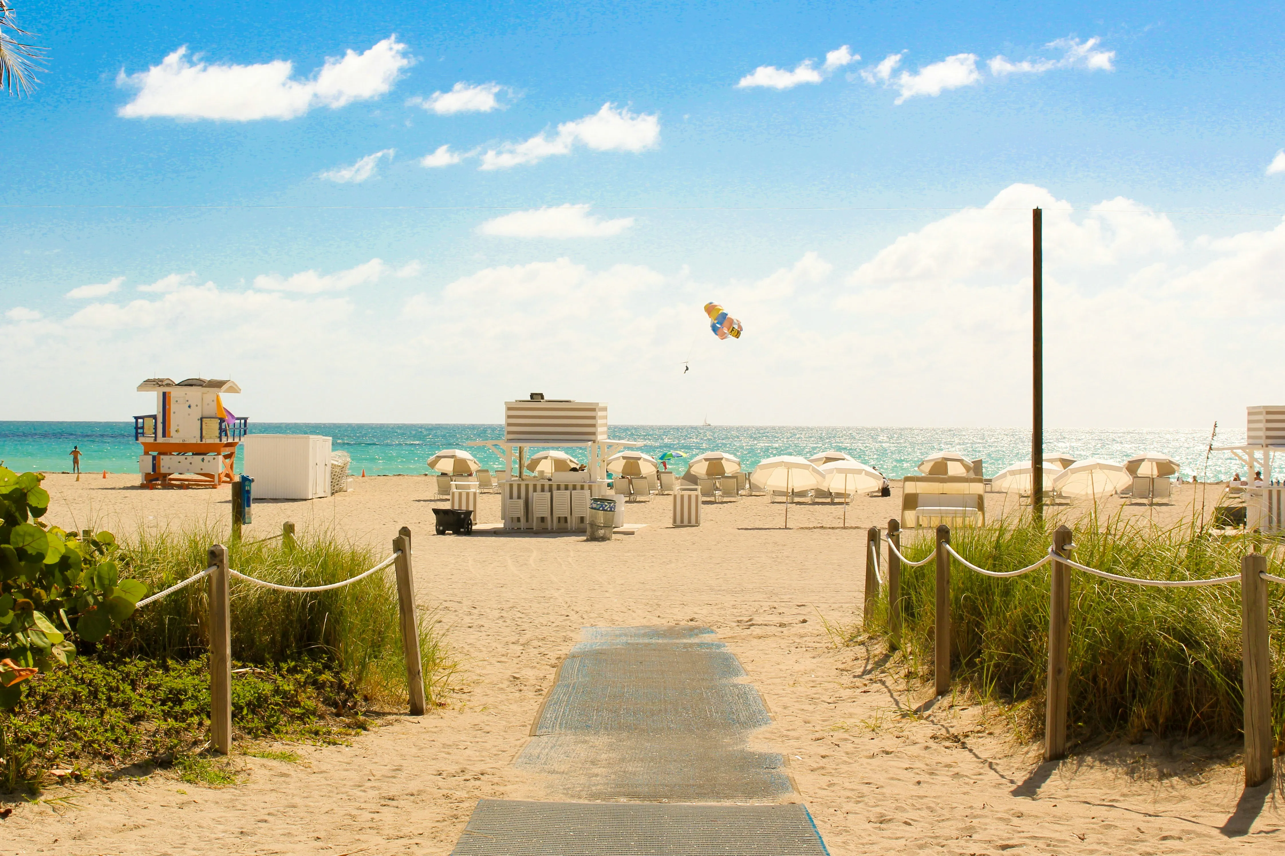 Sandiger Strandzugang mit Sonnenschirmen, Strandhütten und Blick auf das Meer unter blauem Himmel mit wenigen Wolken.