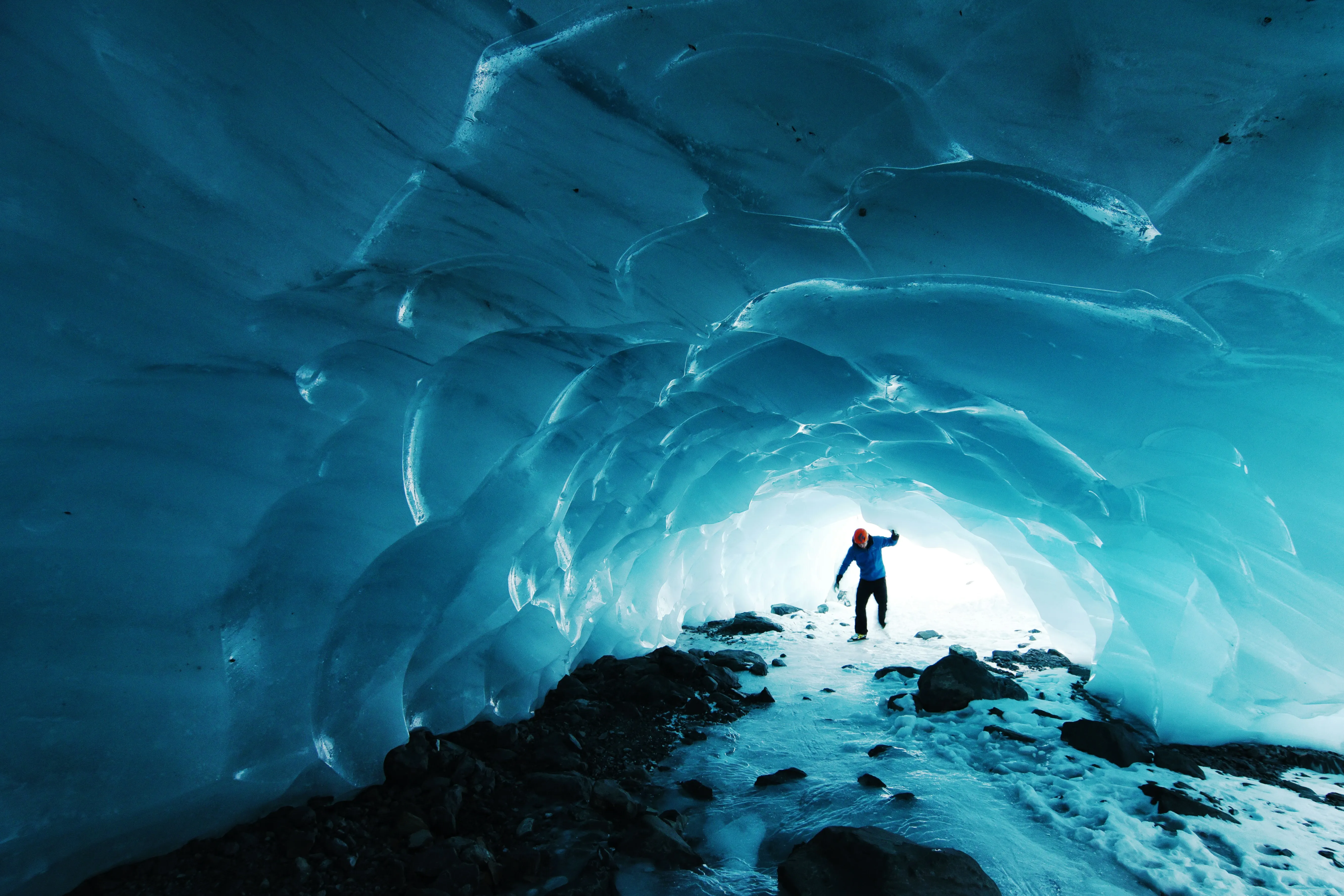 Person in blauer Jacke steht in einem von eisblauen, gewölbten Eisschichten umgebenen Tunnel mit unebenem Boden und Felsen.