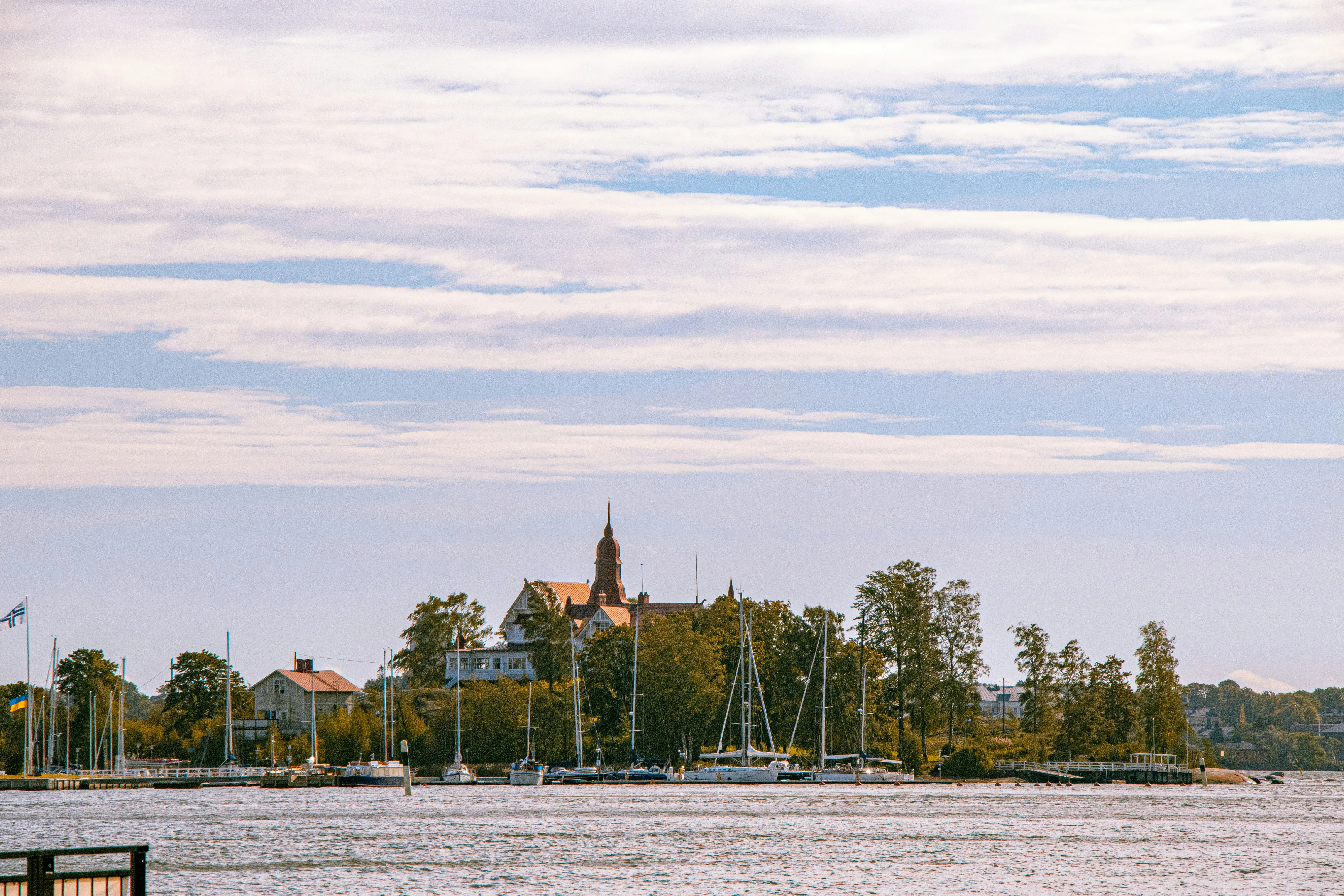 Kleine Insel mit Bäumen, mehreren Segelbooten am Ufer und einem Gebäude mit Turm im Hintergrund unter bewölktem Himmel.