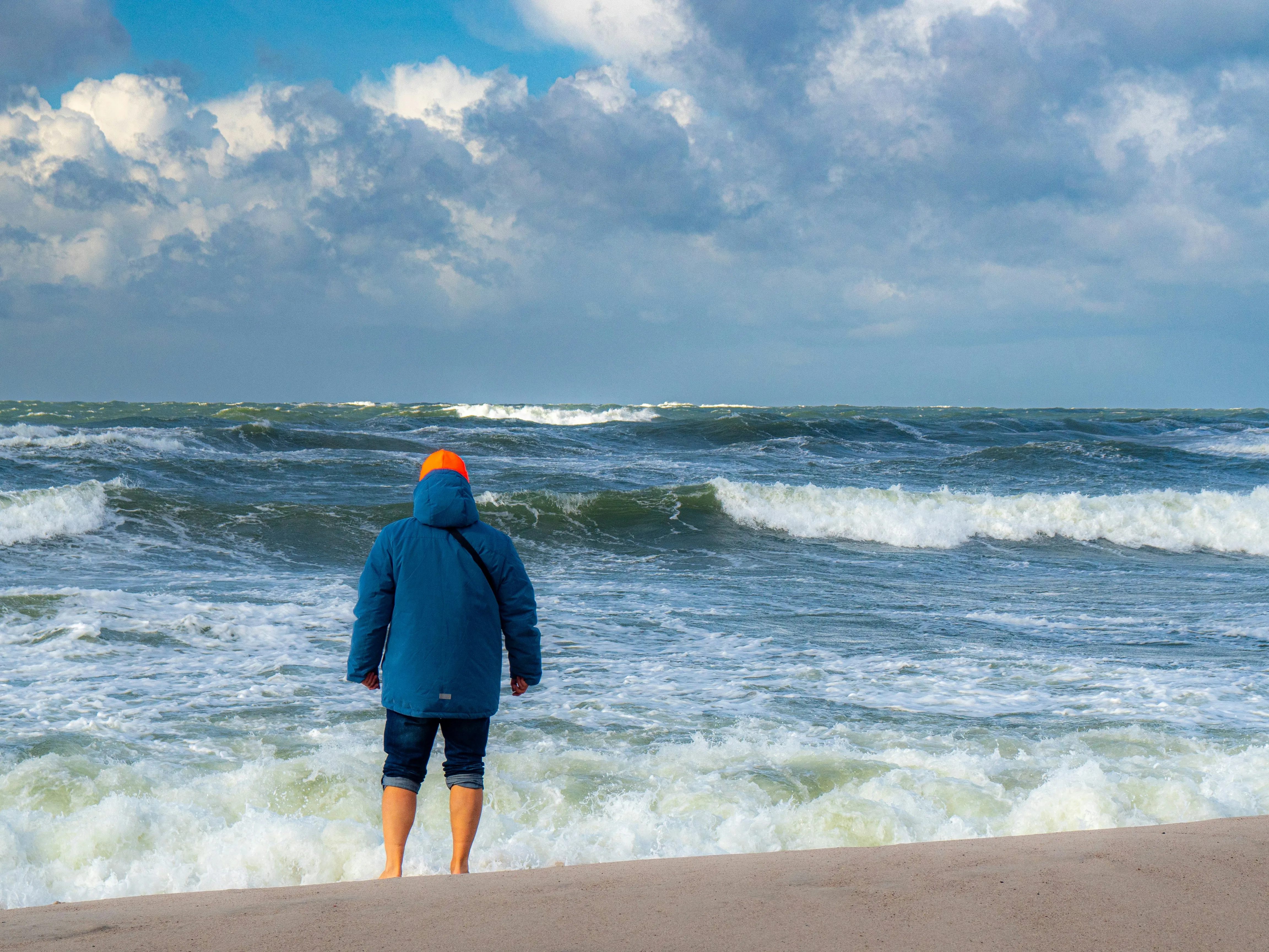 Person mit blauer Jacke und oranger Mütze steht am Strand und blickt auf stürmische See mit hohen Wellen.