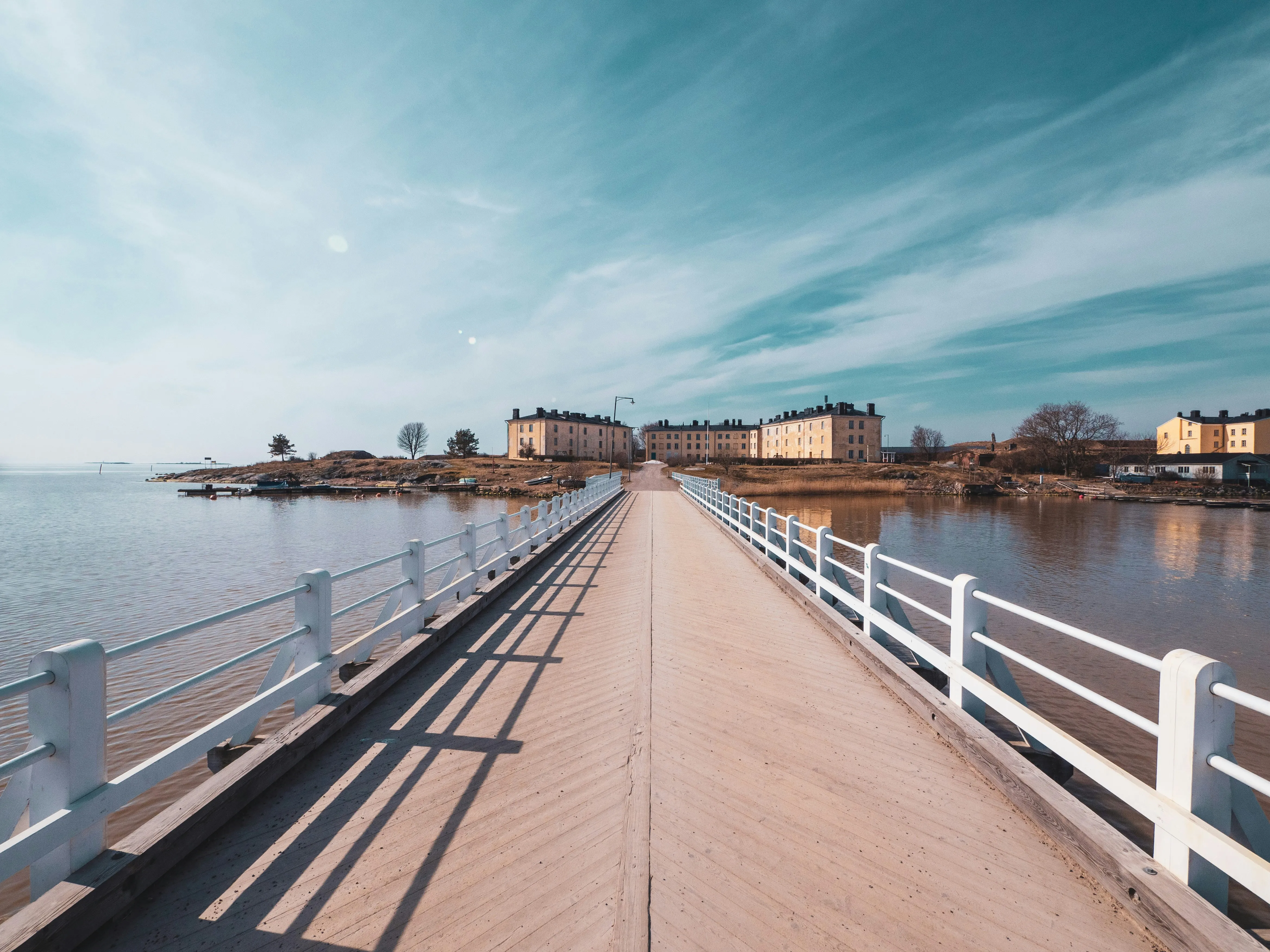 Brücke mit weißen Geländern führt zu mehreren Gebäuden auf einer Insel im Wasser unter klarem Himmel.