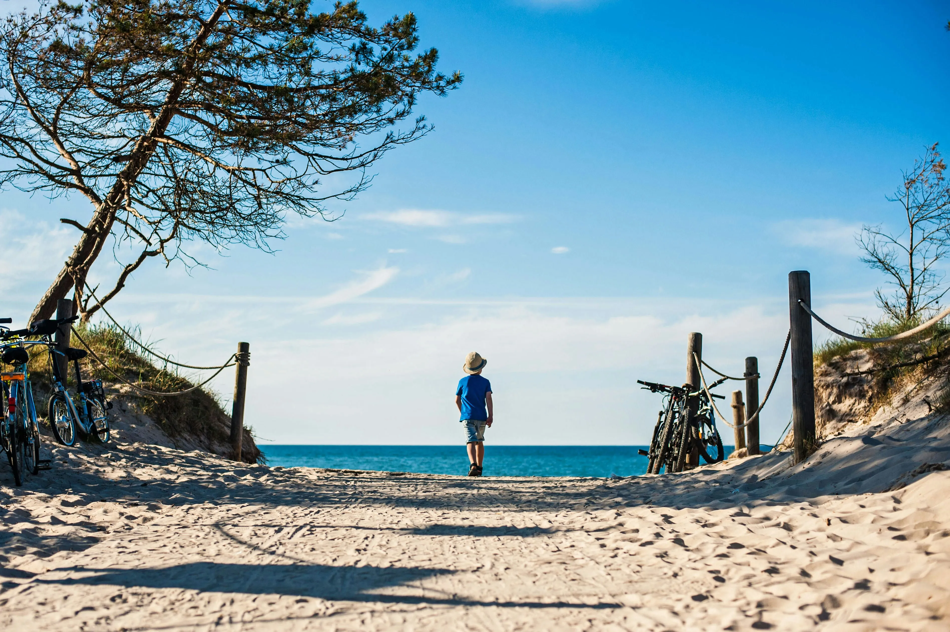 Kind mit Hut läuft auf einem Sandweg zwischen Dünen auf das Meer zu, links und rechts Fahrräder an Pfosten gelehnt.