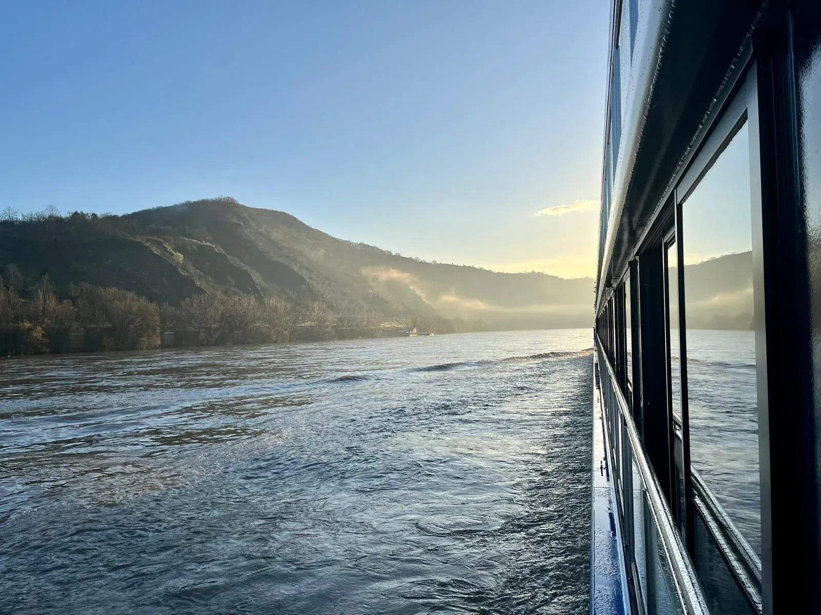 Fluss mit bewaldeten Hügeln im Hintergrund und Spiegelung eines Schiffs an der rechten Bildseite
