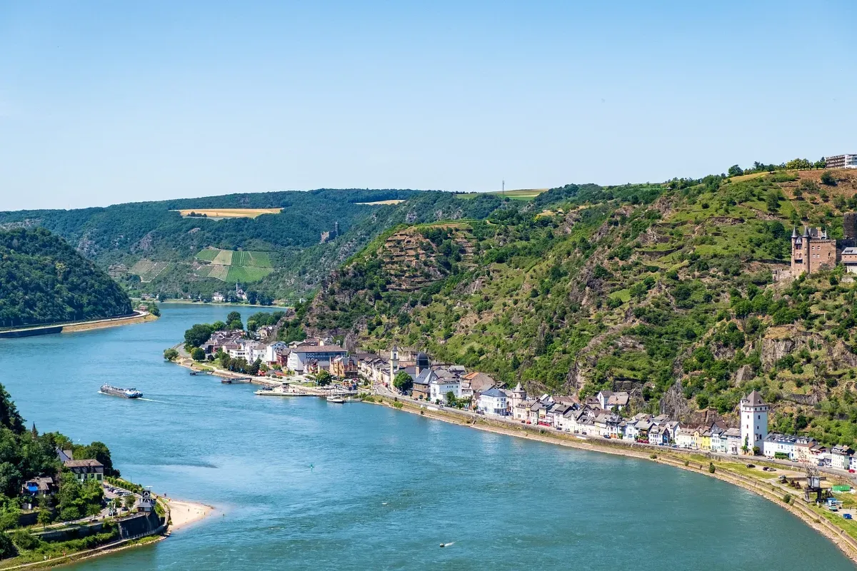 Flusslandschaft am Rhein mit einem breiten Fluss, bewaldeten Hügeln und einem Ort mit Häusern am Ufer