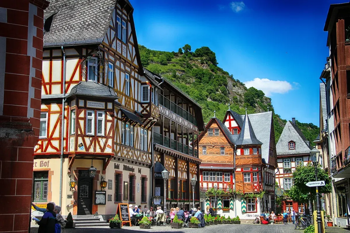 Fachwerkhäuser an einem belebten Platz mit Außengastronomie vor bewaldetem Hügel unter blauem Himmel