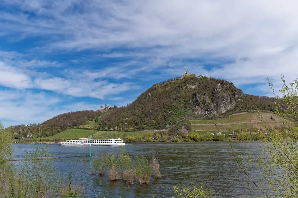 Fluss mit Ausflugsschiff vor bewaldetem Hügel unter blauem Himmel mit Wolken