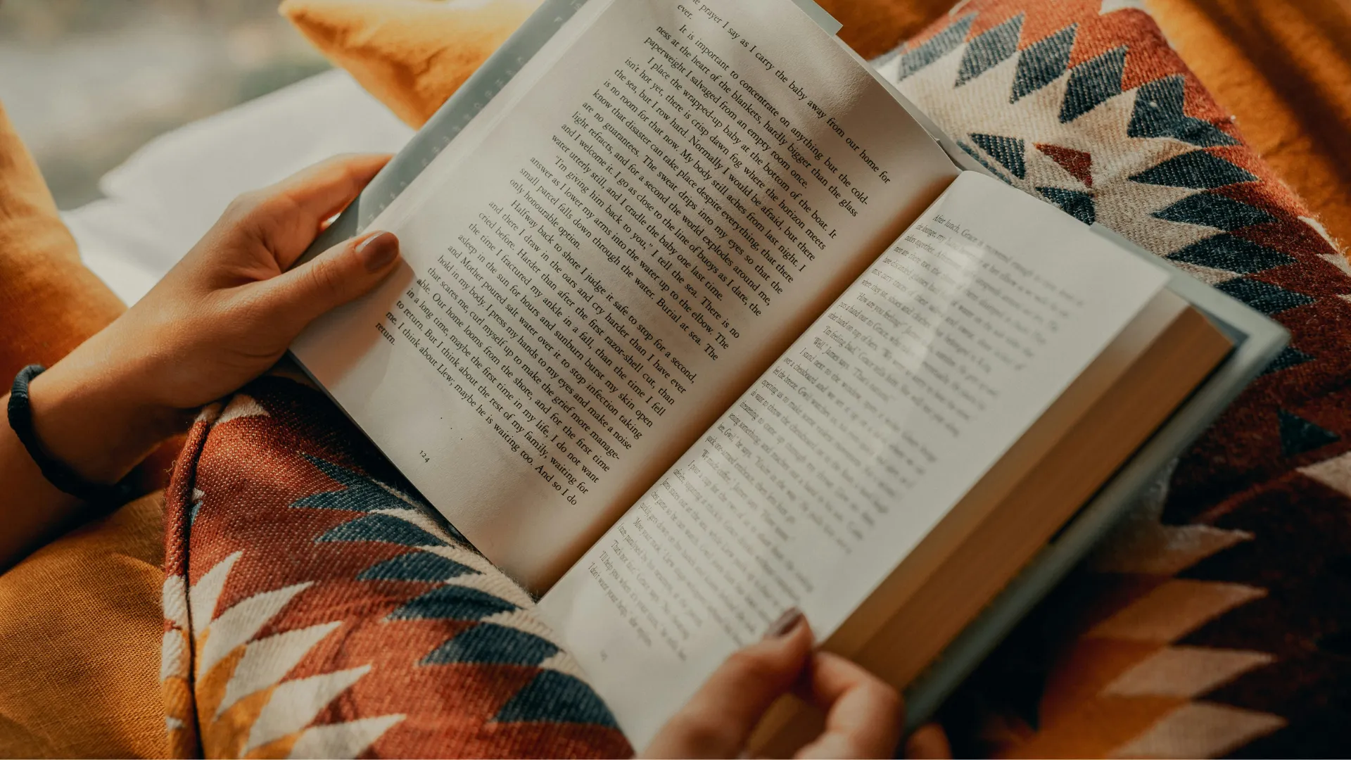 Hände halten ein aufgeschlagenes Buch über einer Decke mit geometrischem Muster.