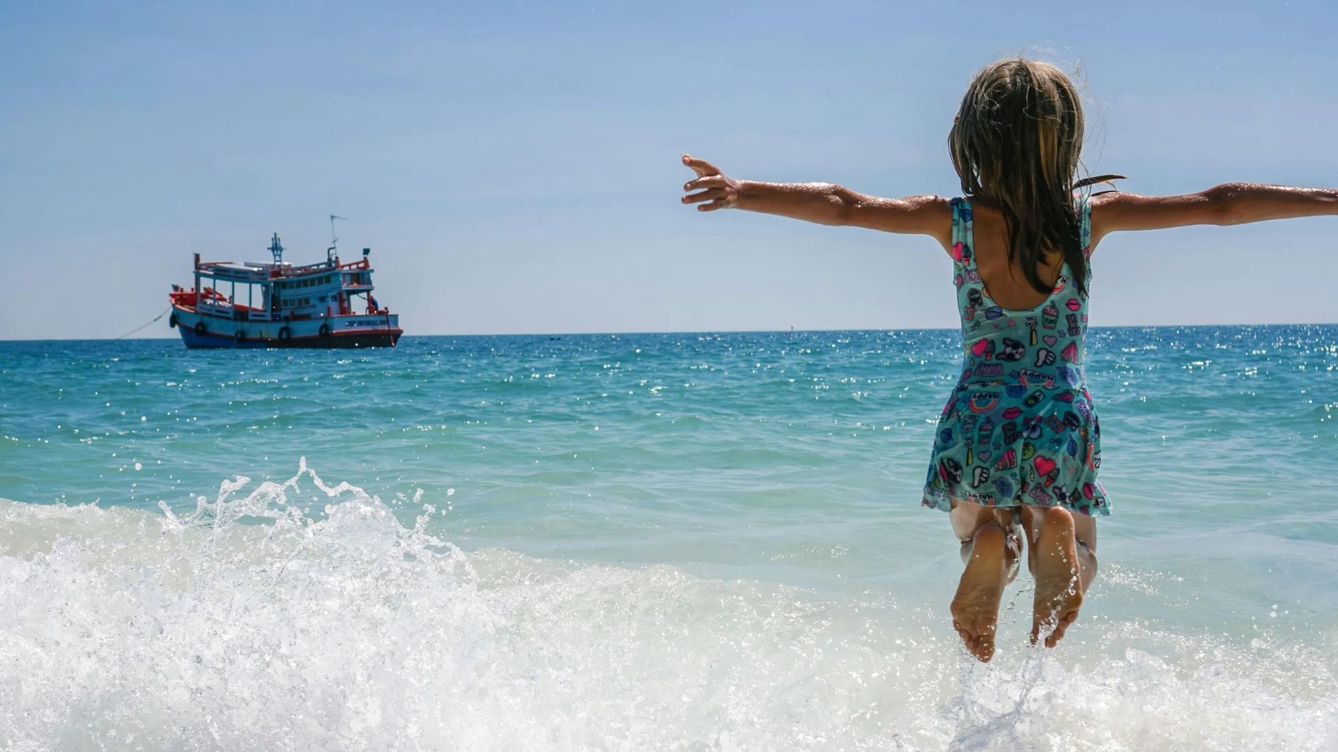 Ein Mädchen im bunten Badeanzug springt mit ausgebreiteten Armen ins Meer, im Hintergrund ein Boot auf dem Wasser.