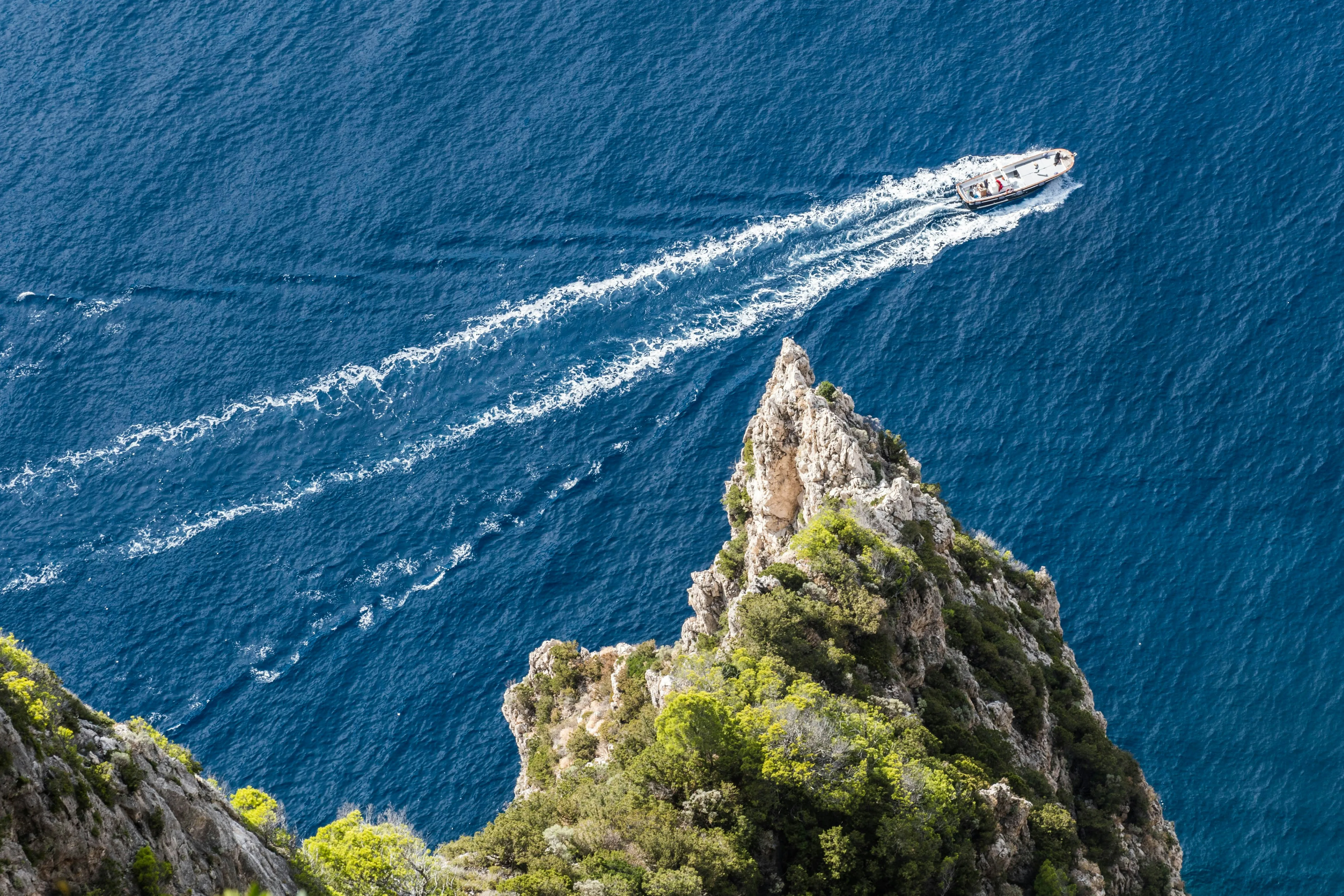 Luftaufnahme eines spitzen Felsvorsprungs mit Vegetation, vor dem ein Motorboot auf blauem Wasser eine weiße Gischtspur zieht.
