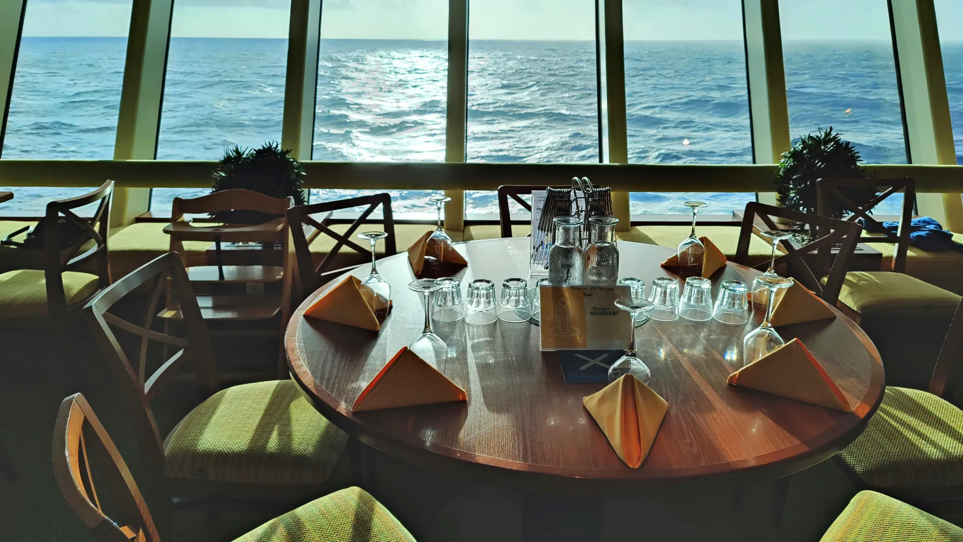 Runder Holztisch mit gefalteten Servietten, Gläsern und Flaschen vor großen Fenstern mit Blick auf das Meer.