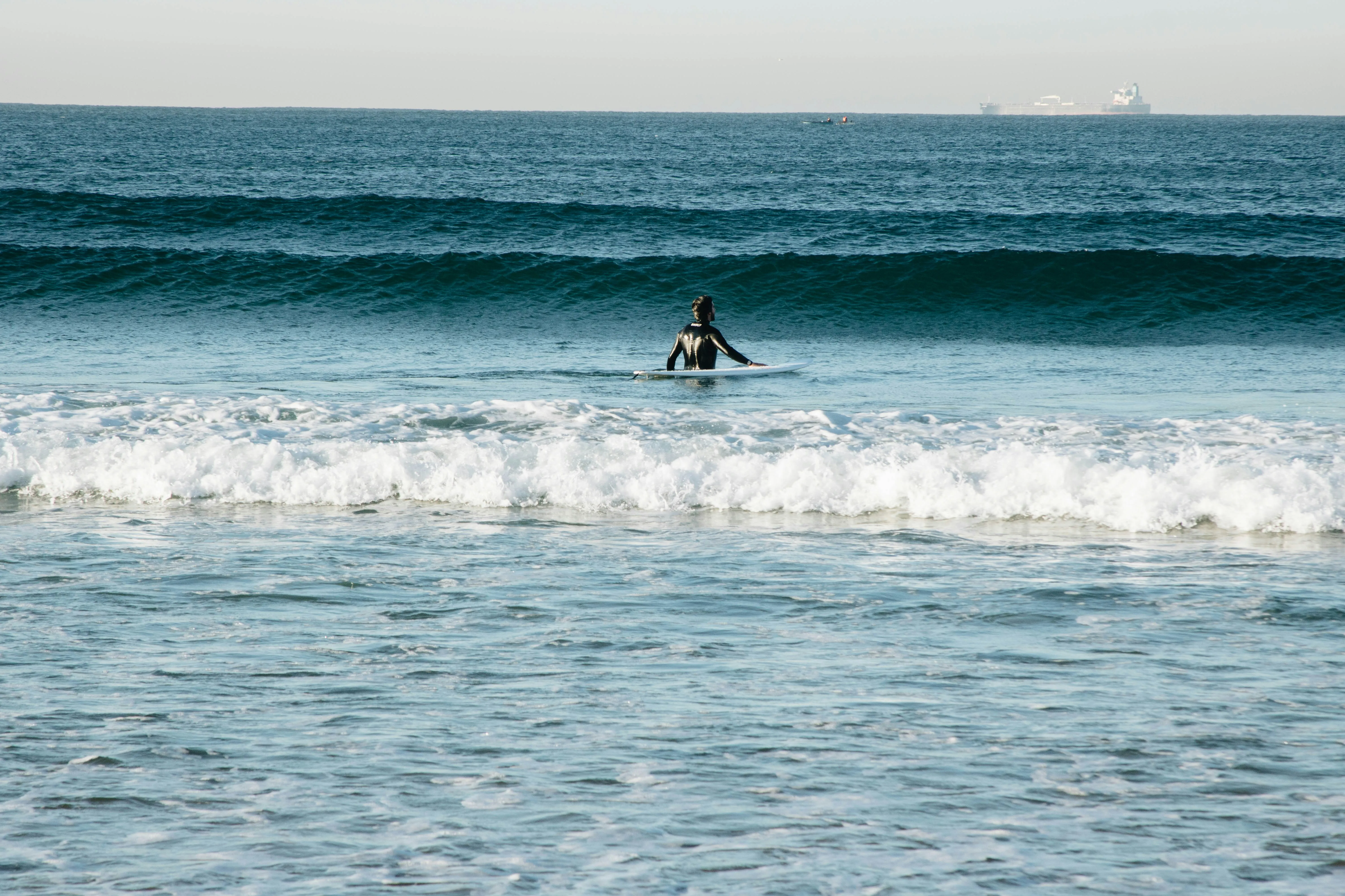 Person in Neoprenanzug sitzt auf Surfbrett im Meer vor einer Welle.