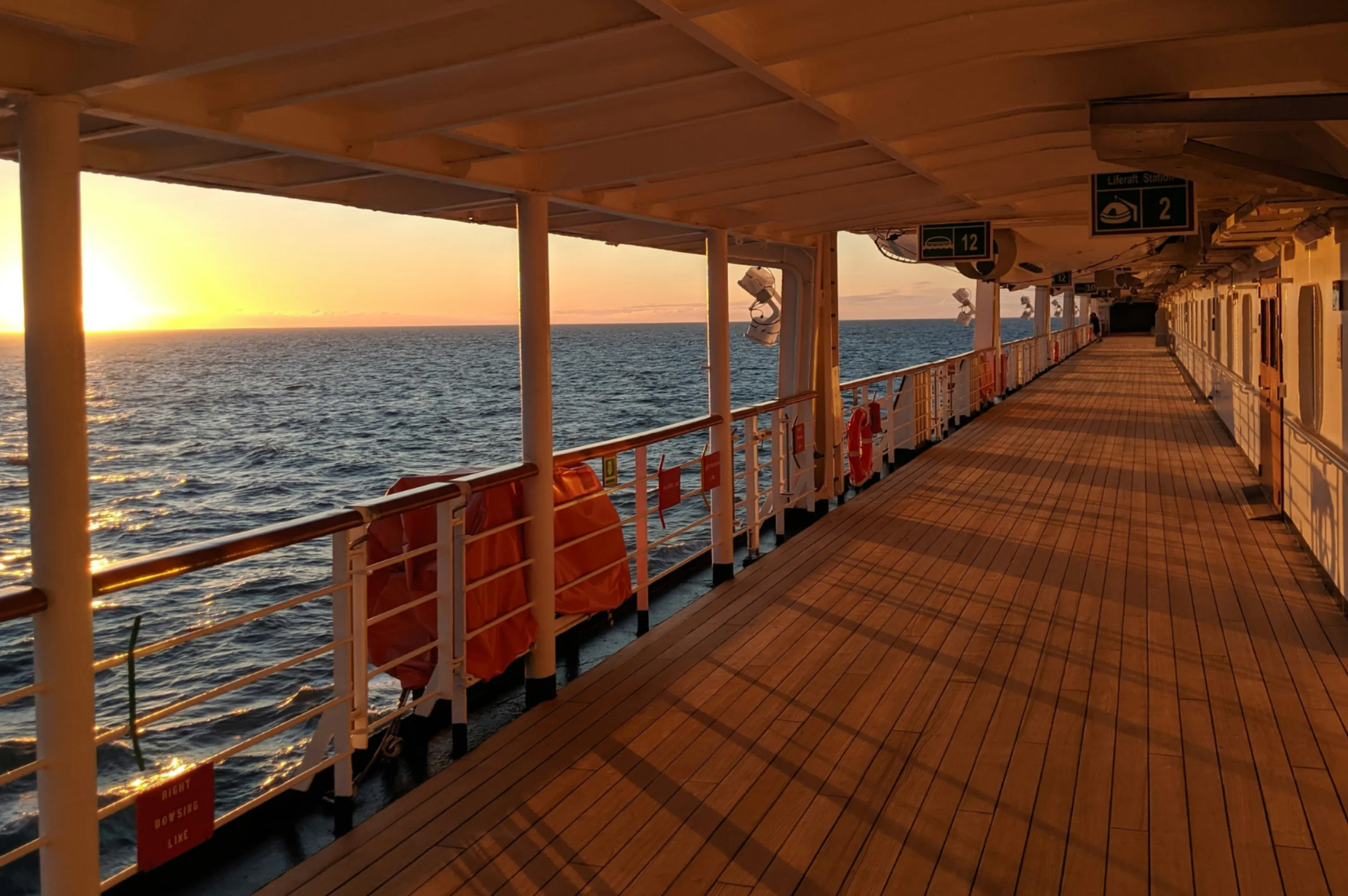 Blick auf das Deck eines Schiffes bei Sonnenuntergang mit Geländern, Rettungsringen und ruhiger See im Hintergrund.