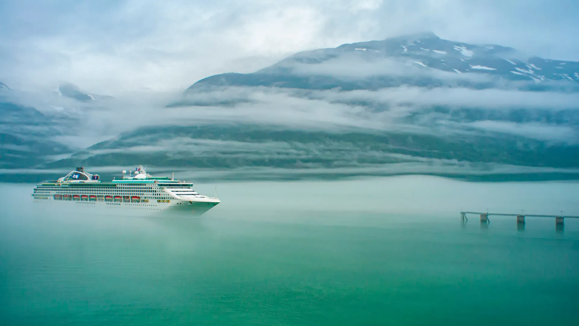 Kreuzfahrtschiff auf ruhigem Wasser vor nebelverhüllten Bergen mit schneebedeckten Gipfeln.