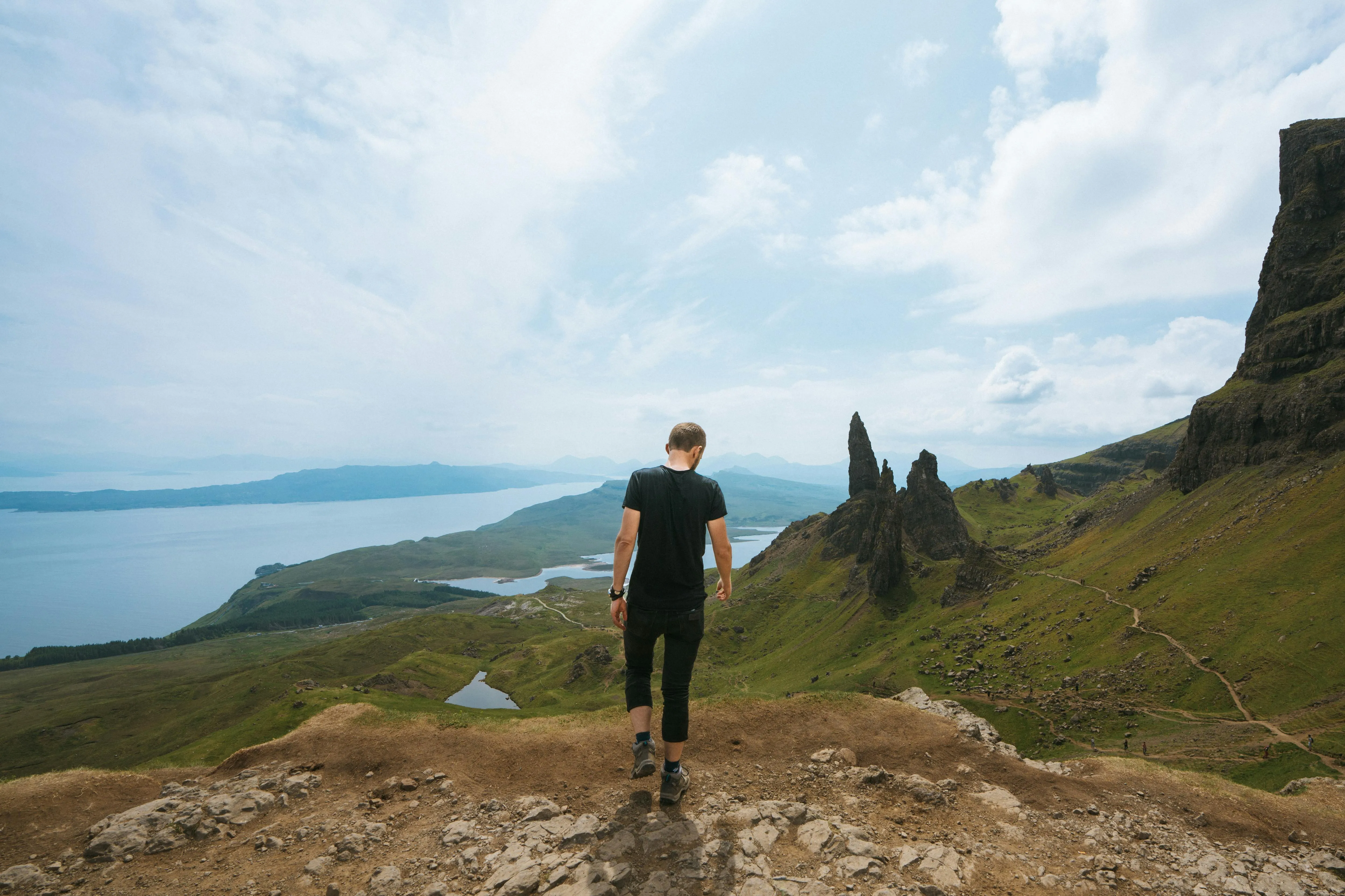 Person in schwarzem T-Shirt und Hose wandert auf einem felsigen Pfad in einer grünen Berglandschaft mit markanten Felsnadeln und weiter Sicht auf Wasser und Himmel.