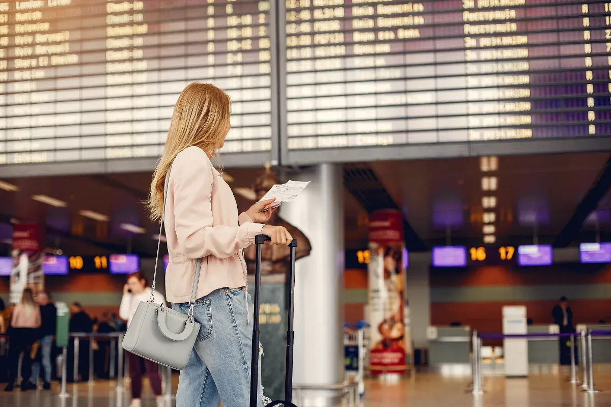 Frau mit langem Haar, rosa Jacke und Jeans steht mit Rollkoffer und Flugticket vor einer großen Anzeigetafel im Flughafen