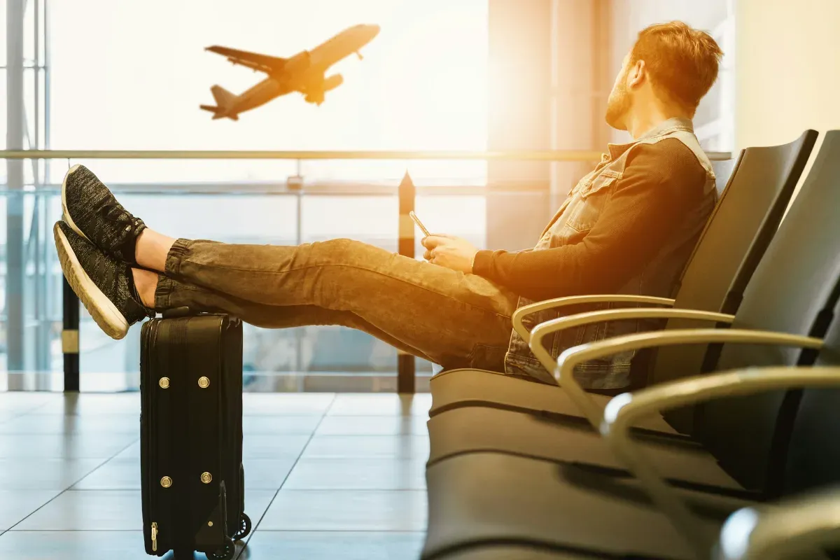 Mann sitzt mit ausgestreckten Beinen auf einem Stuhl im Flughafen und blickt auf startendes Flugzeug