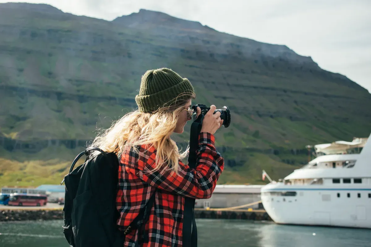 Frau mit grüner Mütze und rotschwarz kariertem Hemd fotografiert vor einem Kreuzfahrtschiff in einem Fjord