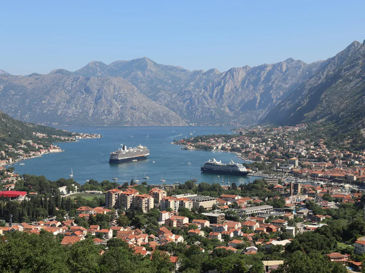 Zwei Kreuzfahrtschiffe liegen in einem Hafen an, im Vordergrund ist eine Stadt mit vielen roten Dächern zu sehen und in der Umgebung viele Berge