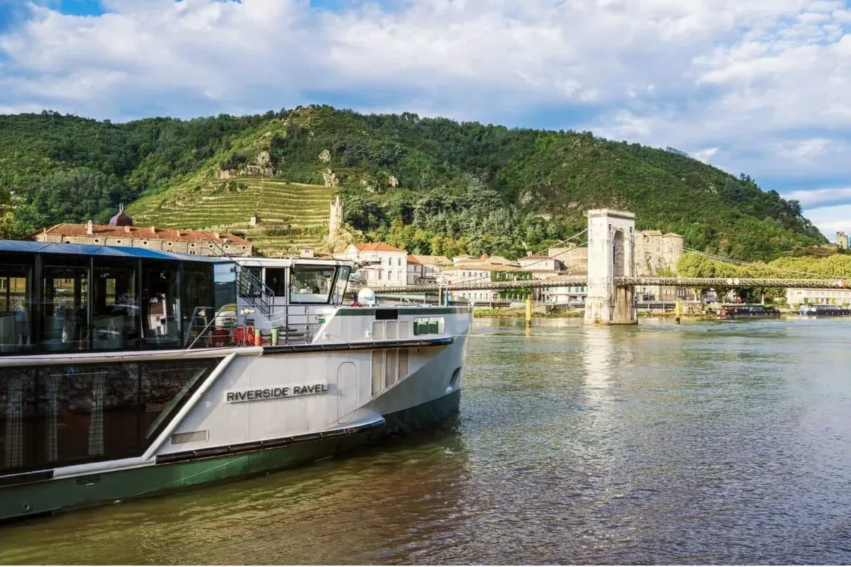 Flussufer mit Ausflugsschiff namens 'RIVERSIDE RAVEL' im Vordergrund und bewaldetem Hügel mit Weinbergen im Hintergrund