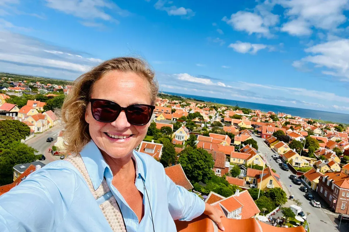 Frau mit hellblondem lockigem Haar in hellblauem Hemd macht Selfie mit Blick auf ein Dorf mit roten Dächern und Meer im Hintergrund