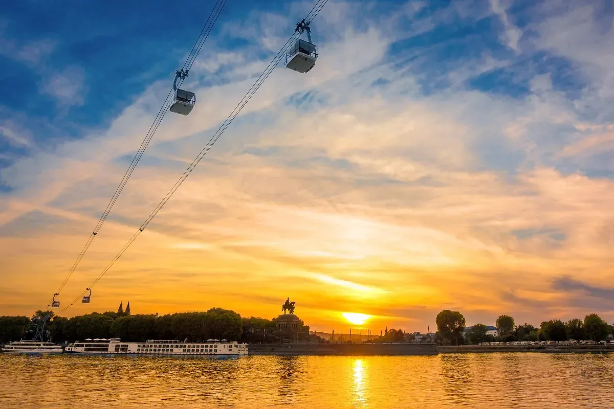 Seilbahnkabinen über einem Fluss bei Sonnenuntergang mit einer Statue und Bäumen am Ufer