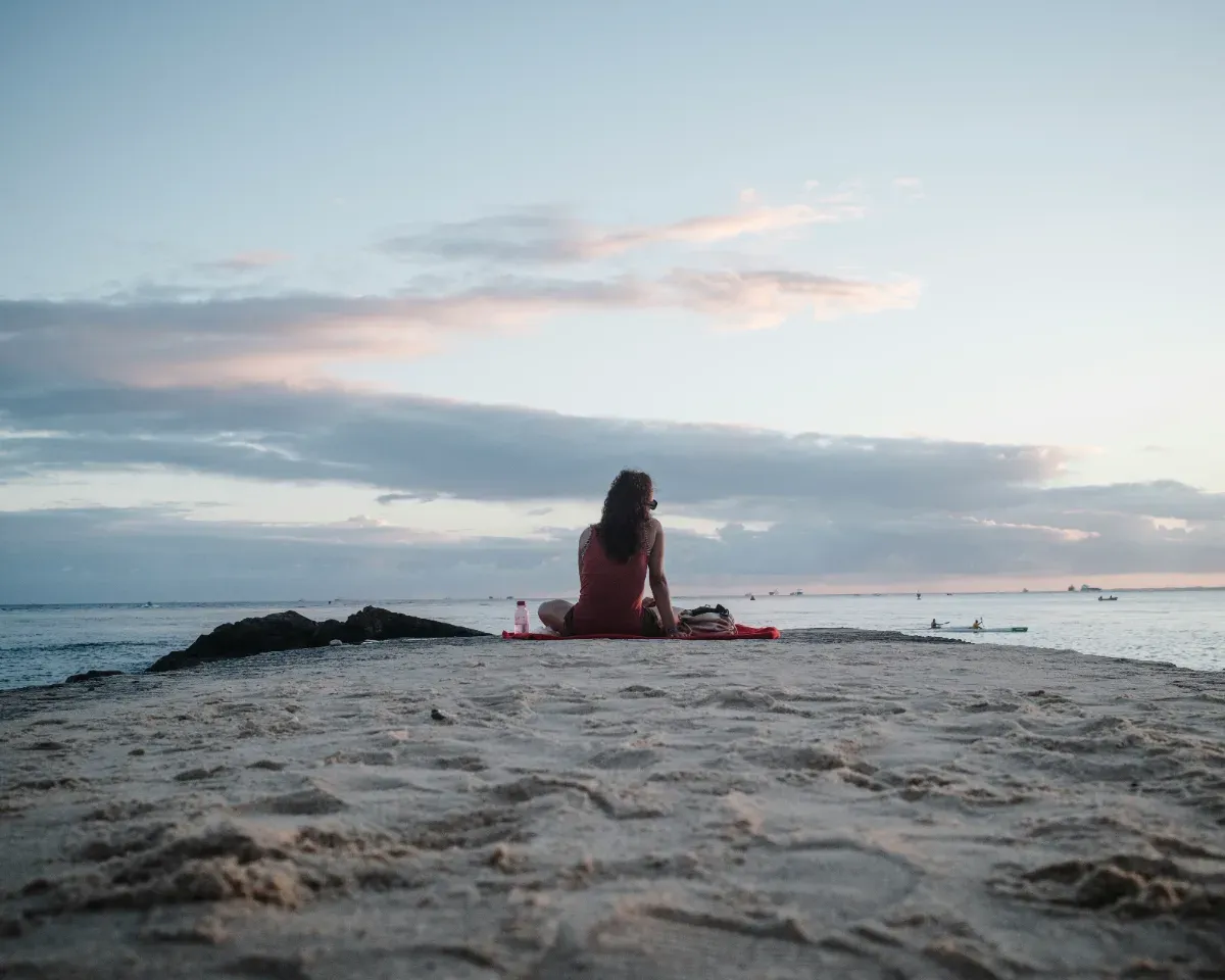 Person sitzt auf einem Sandweg am Meer und blickt auf den Horizont bei bewölktem Himmel
