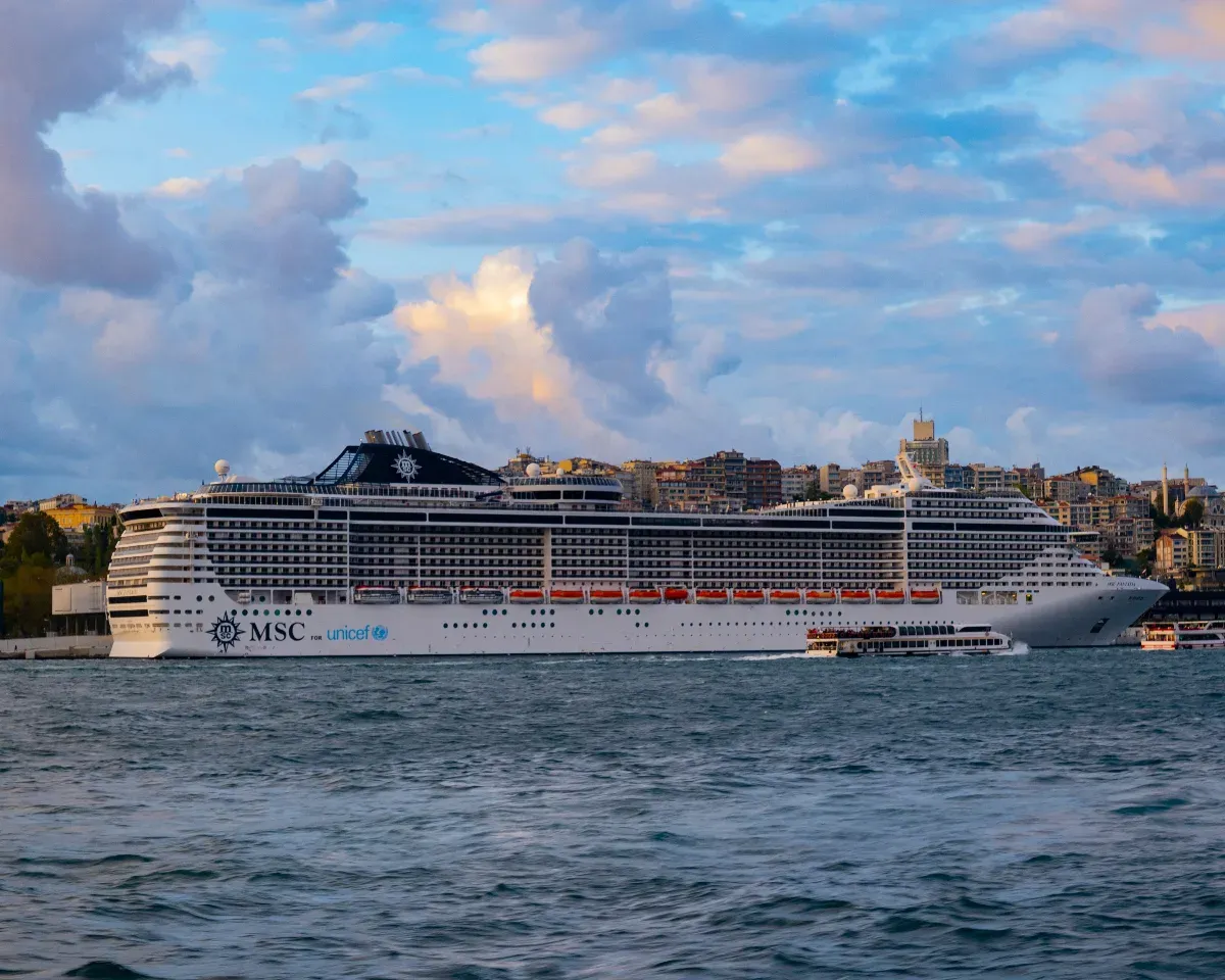 Großes Kreuzfahrtschiff der Marke MSC auf ruhigem Wasser vor einer Stadt mit mehrstöckigen Gebäuden unter bewölktem Himmel