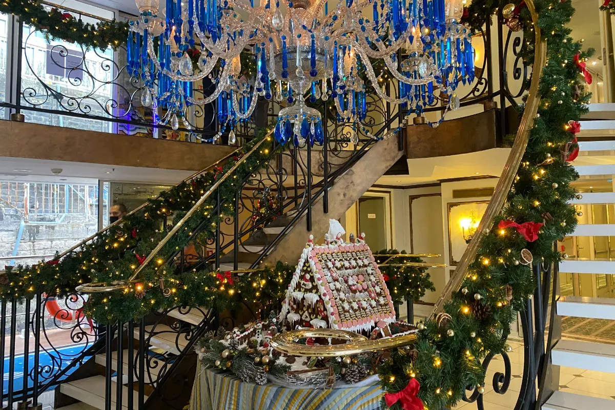 Weihnachtlich geschmücktes Treppengeländer mit Lichterketten und roten Schleifen in einem Innenraum mit blau-weißer Kristallleuchterdecke