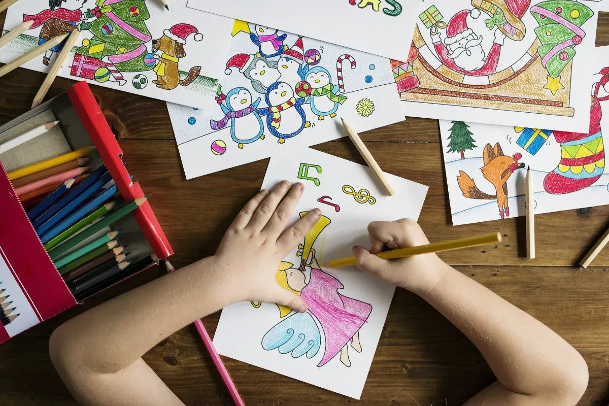 Kinderhände malen mit Buntstiften bunte Weihnachtsmotive auf Papier, darunter Engel, Pinguine und Weihnachtsmann