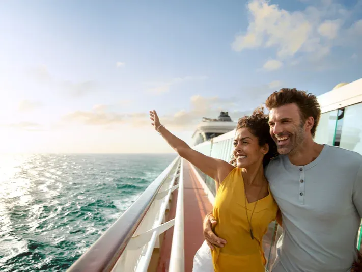 Paar steht auf dem Deck eines Kreuzfahrtschiffs und blickt aufs Meer, Frau hebt den Arm