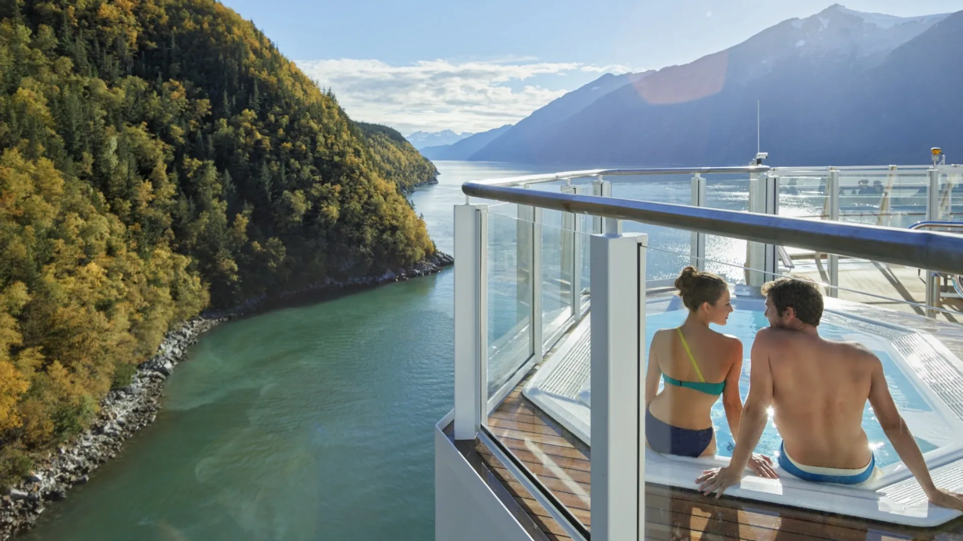 Zwei Personen sitzen in einem Whirlpool auf einem Schiff mit Blick auf einen Fluss und bewaldete Berge