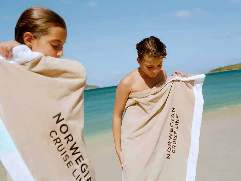 Zwei Personen am Strand, die jeweils ein großes Handtuch mit der Aufschrift 'NORWEGIAN CRUISE LINE' um den Körper gewickelt haben